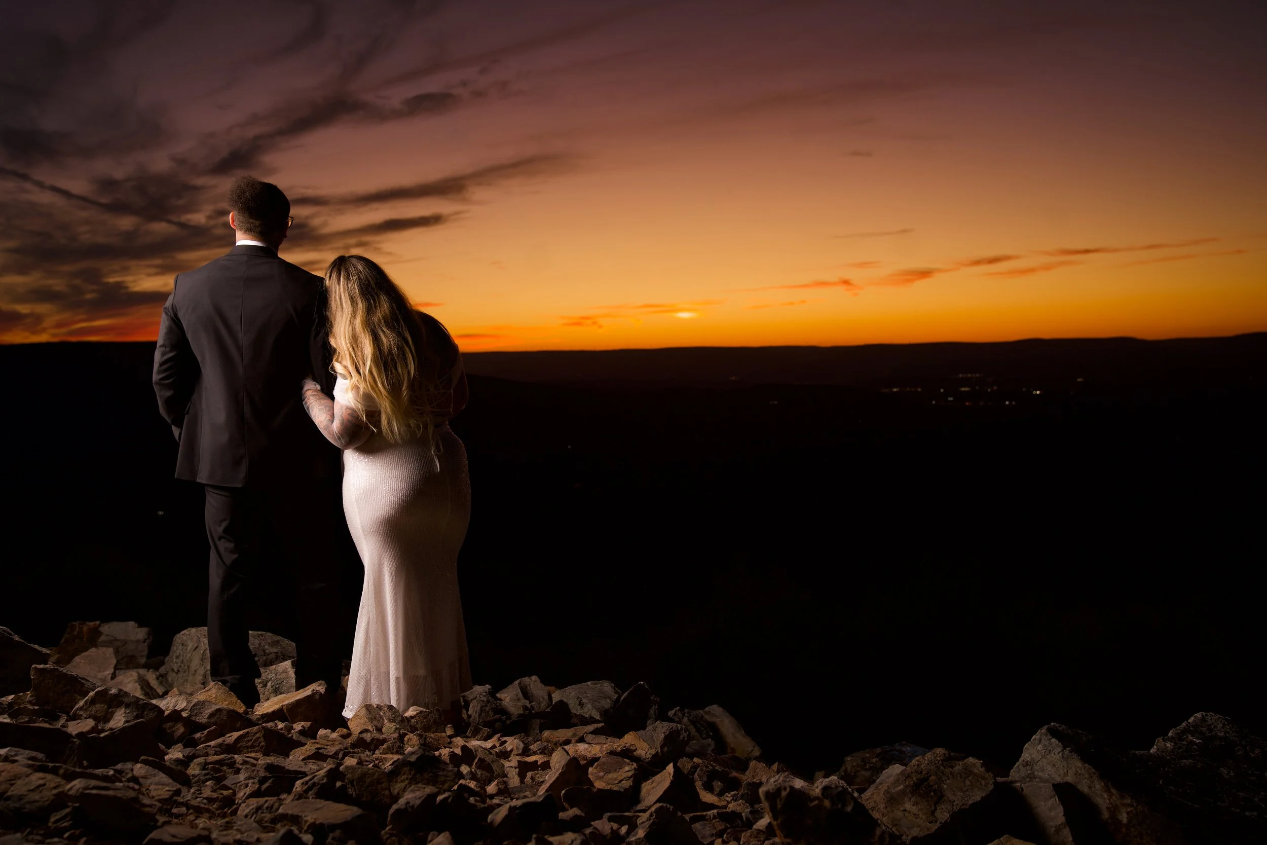 A couple dressed formally, the man in a suit and the woman in a long dress, standing on rocks and looking at a sunset over a landscape.