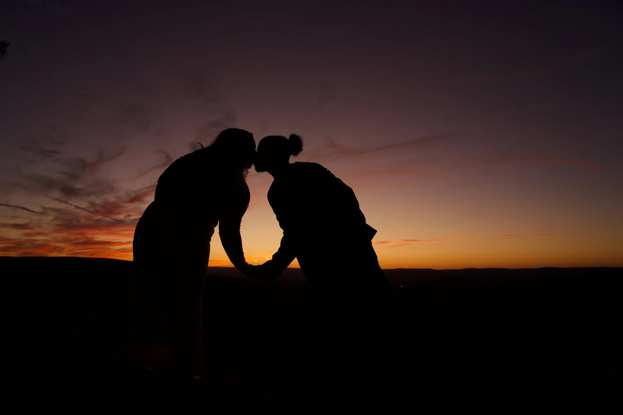 Silhouette of two people holding hands and kissing at sunset on a hill.