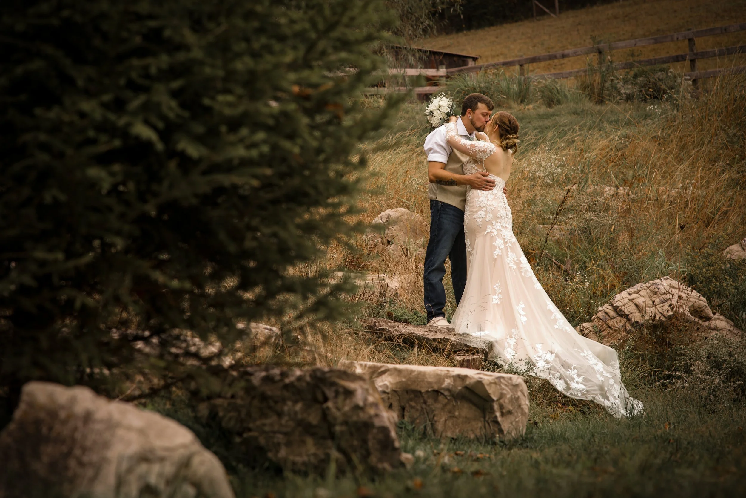 A bride and groom sharing a kiss outdoors on a rocky hillside, with the bride in a white lace wedding gown and the groom in casual attire. The scene is surrounded by natural scenery including rocks, grass, and trees.