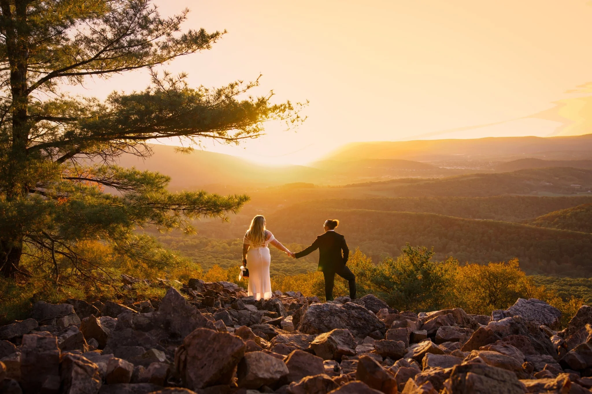 A couple, dressed in formal attire, holds hands on a rocky hilltop during sunset with a scenic view of rolling hills and trees in the background.