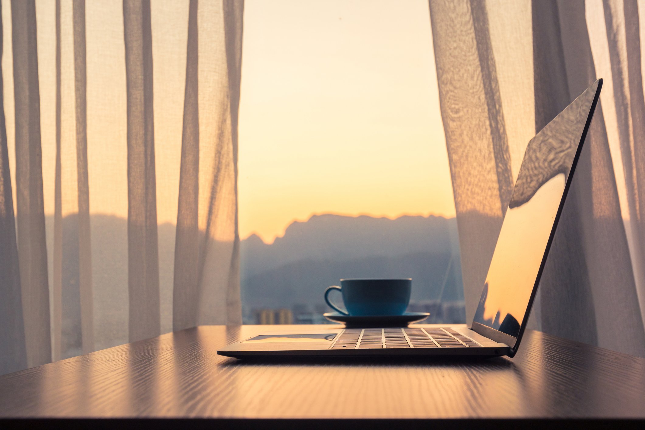 A laptop and a blue cup on a table with sheer curtains and a sunset view of mountains in the background.