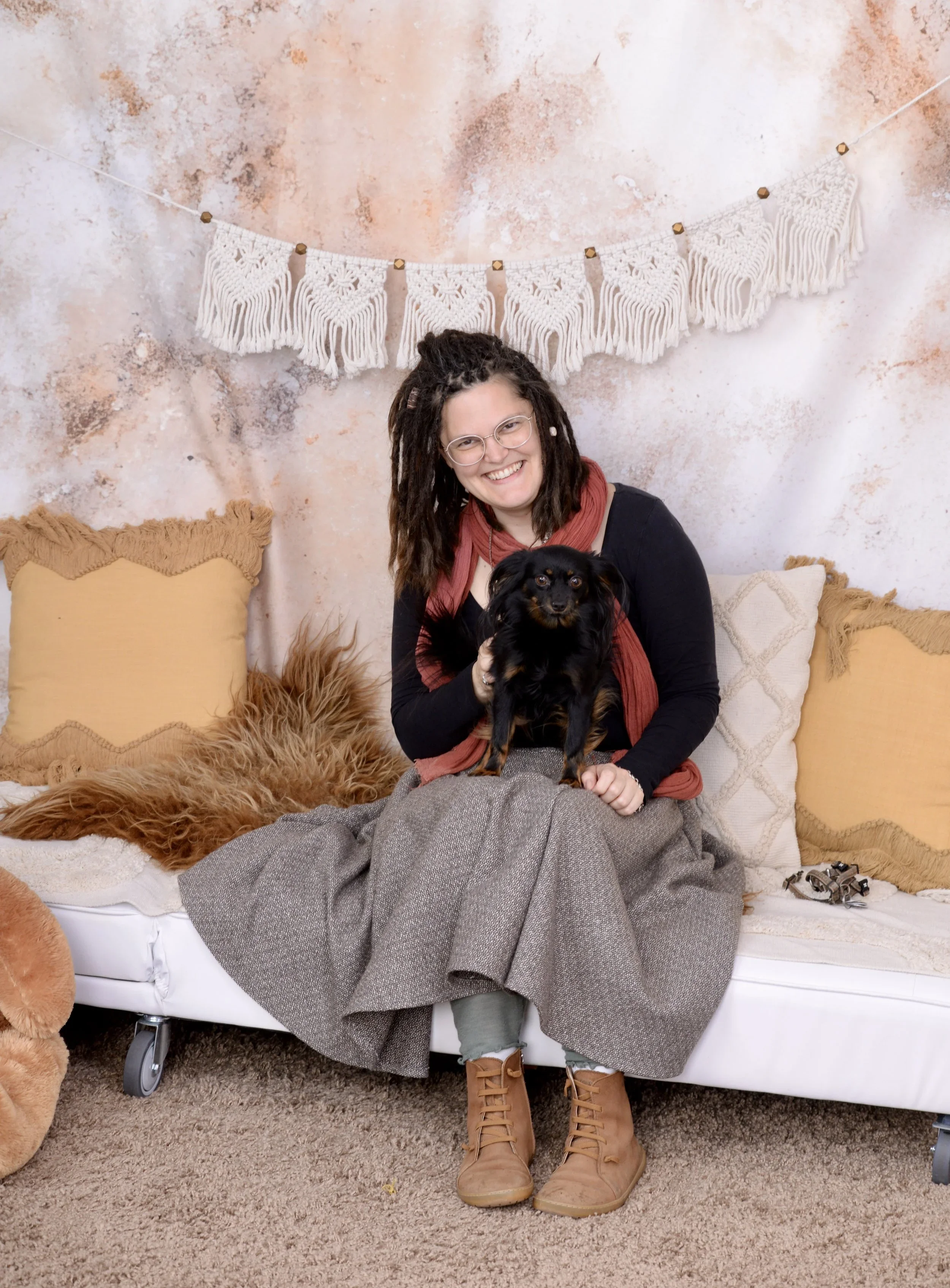 Eine lachende Frau sitzt auf einer Couch mit einem kleinen schwarzen Hund auf ihrem Schoß. Im Hintergrund sind Kissen, ein Fell und eine Wanddekoration aus Makramee sichtbar.