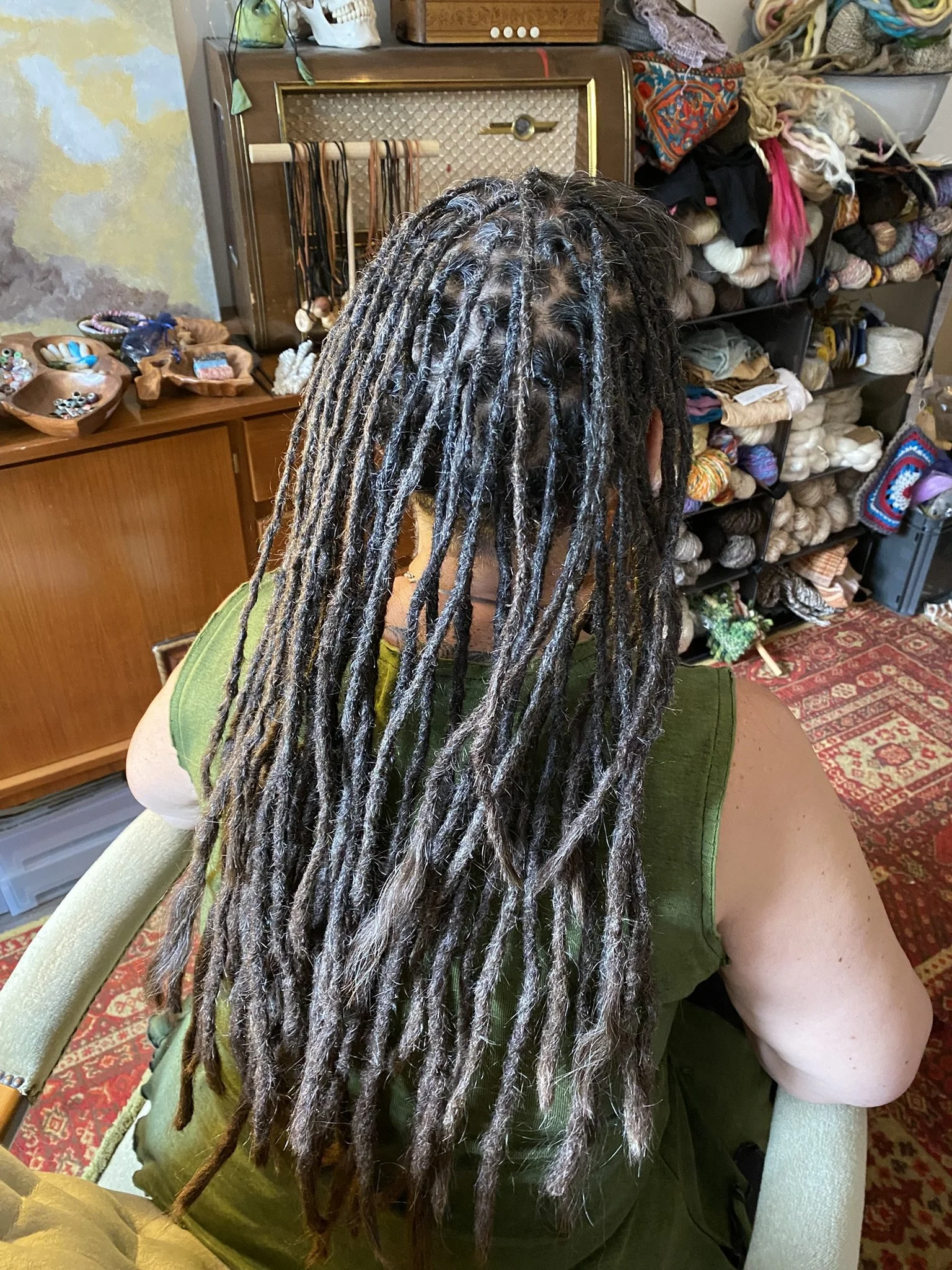 Eine Person mit langem, grau-schwarz gefärbtem Dreadlocks-Haar sitzt in einem Raum mit bunten Wollknäueln und Häkelzubehör im Hintergrund.