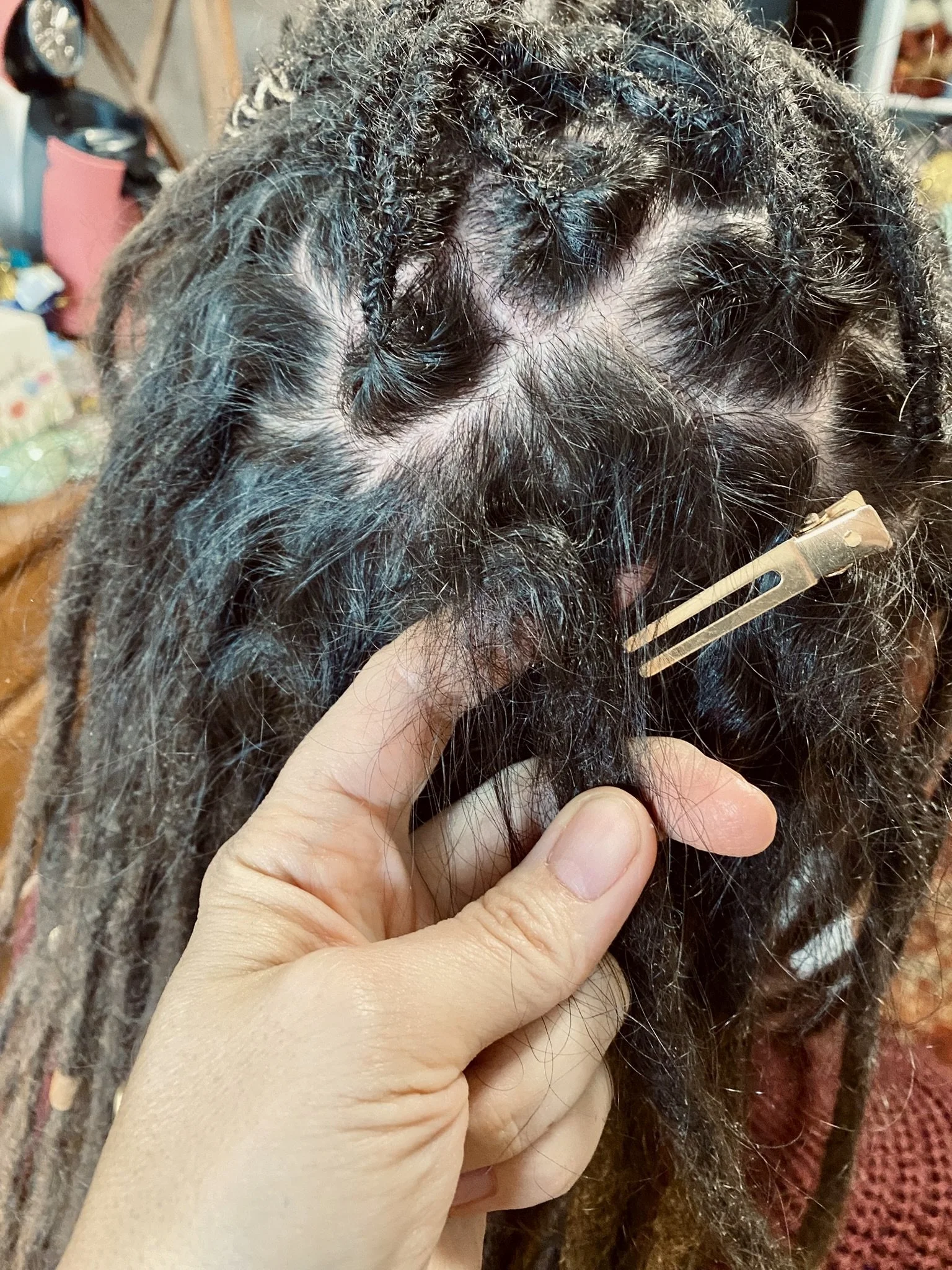 Händliche Person hält Dreadlocks fest, die an einem Kopf mit Zipfelmütze befestigt sind, mit einer Haarklammer. Hintergrund ist ein Raum mit Haushaltsgegenständen.