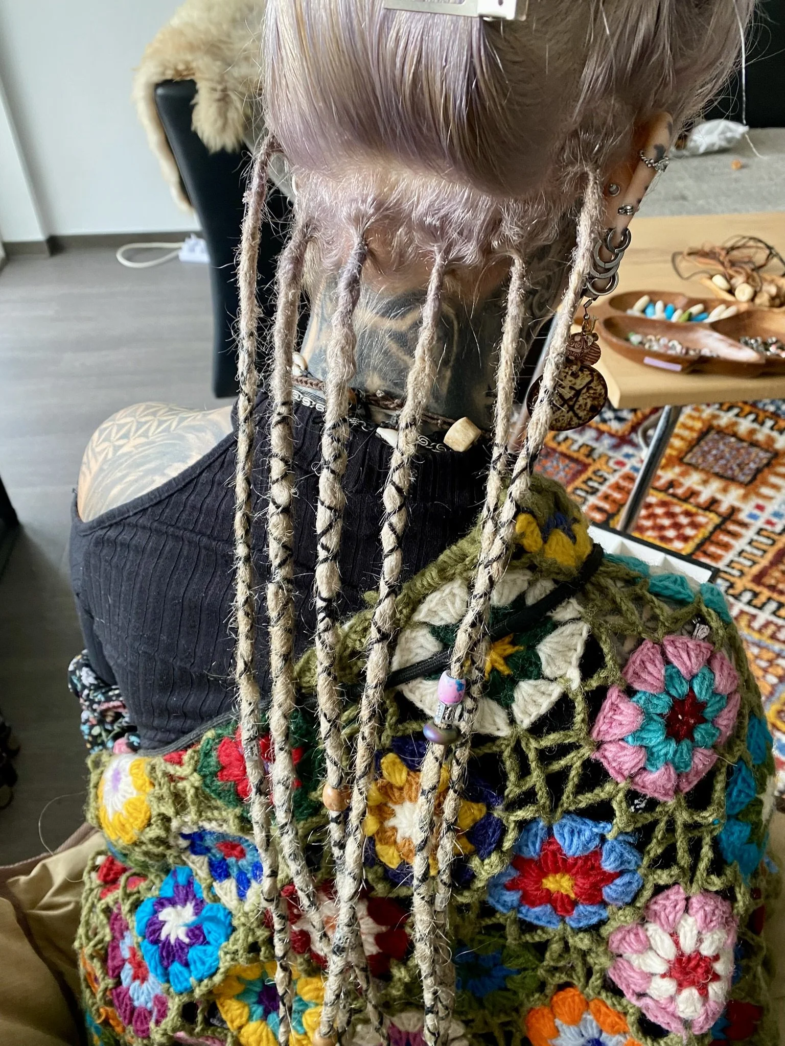 Eine Person mit langen, geflochtenen Haaren und mehreren Piercings trägt eine farbenfrohe, gehäkelte Decke. Im Hintergrund sind Tische mit Schmuck und anderen Gegenständen zu sehen.