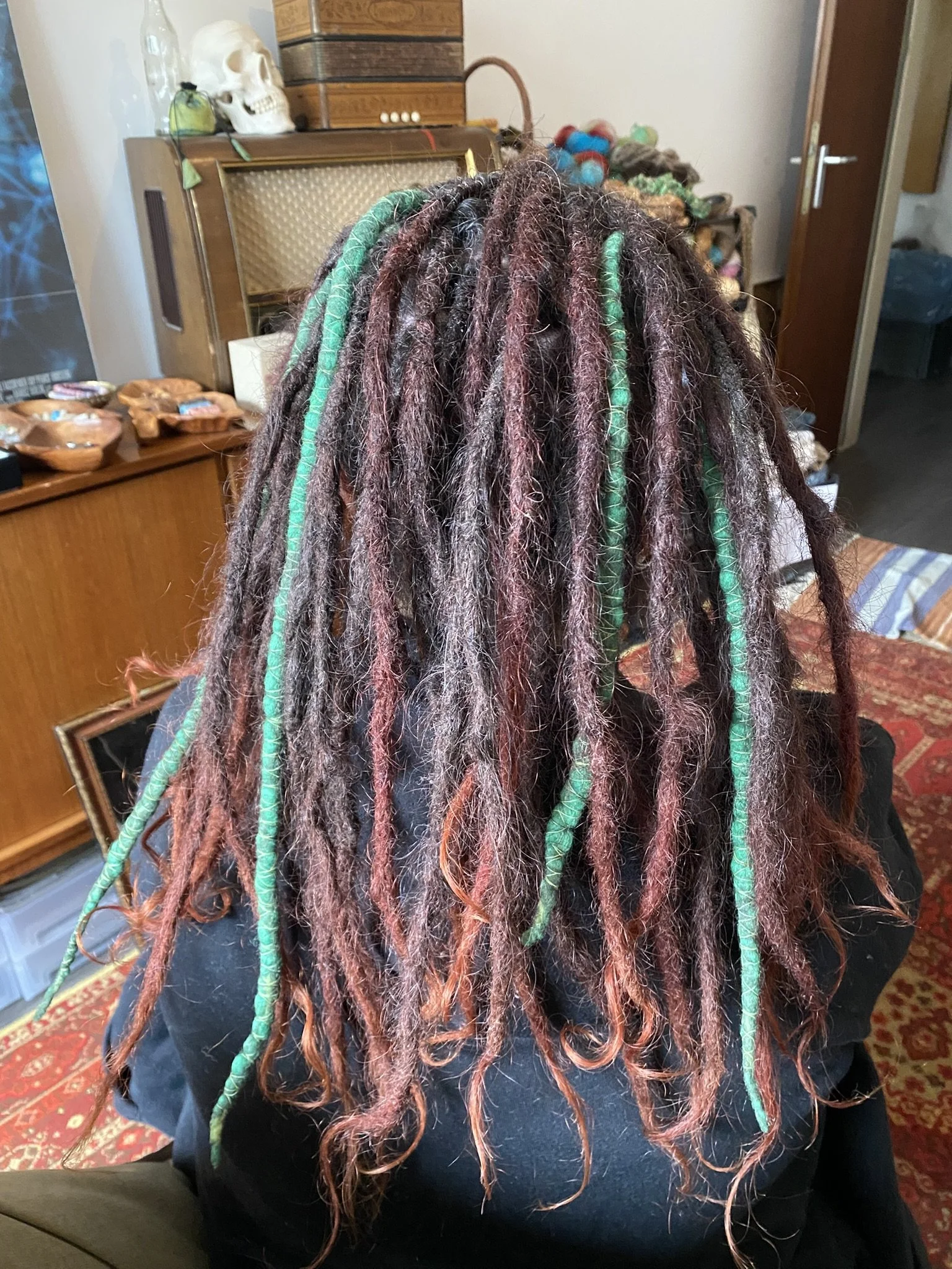 Dreadlocks mit bunten grünen und braunen Neuschichten, auf einem Rücken sitzend, in einem Raum mit Holzmöbeln, Dekorationen und einem Teppich.