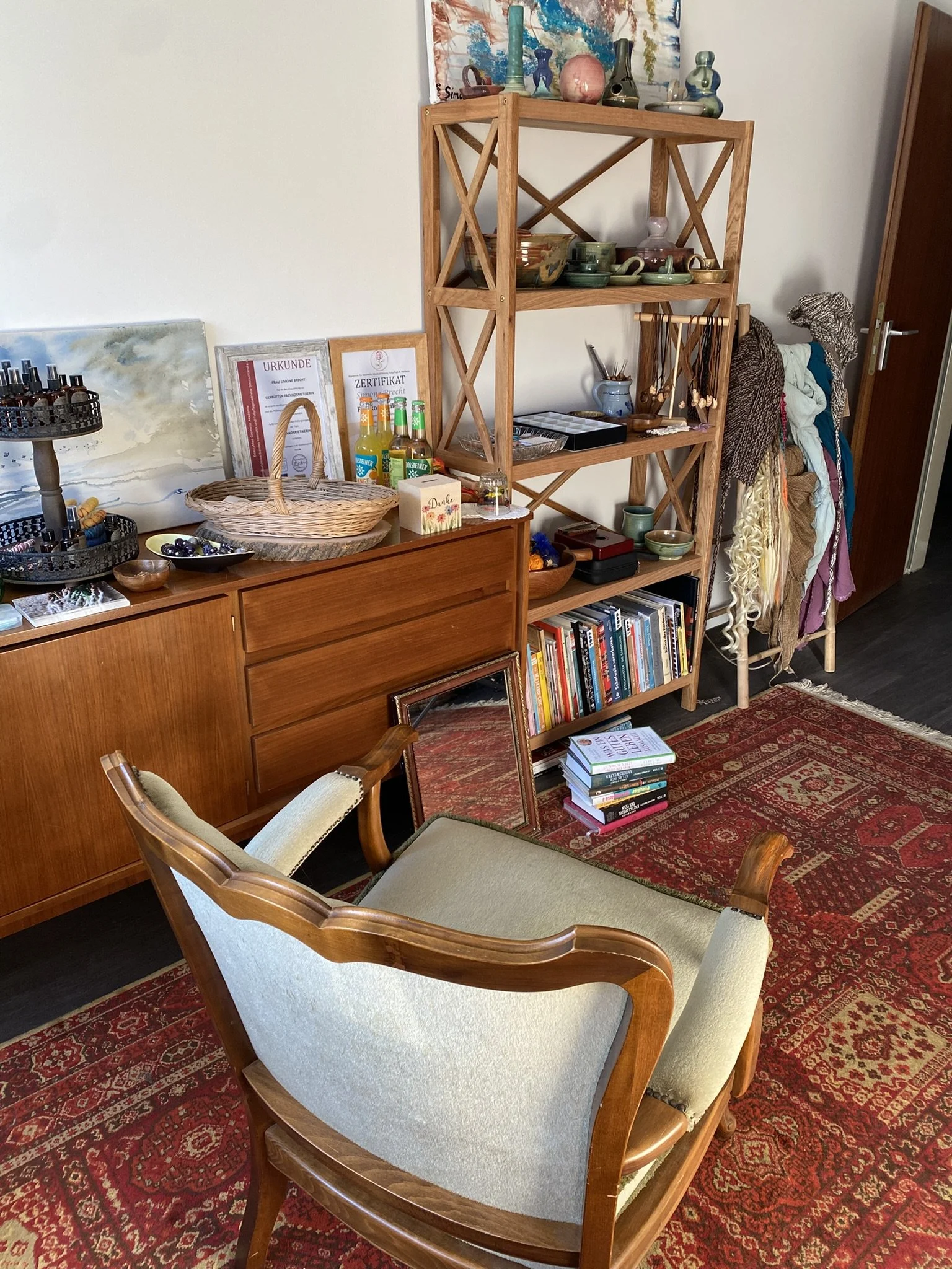 Eine Wohnzimmer-Ecke mit einem alten Holztisch, einem beigen und grünen Armstuhl, einem Holzregal mit Geschirr, Büchern und Dekorationsgegenständen, einem roten Teppich und einigen zerstreuten Büchern und Bildern.