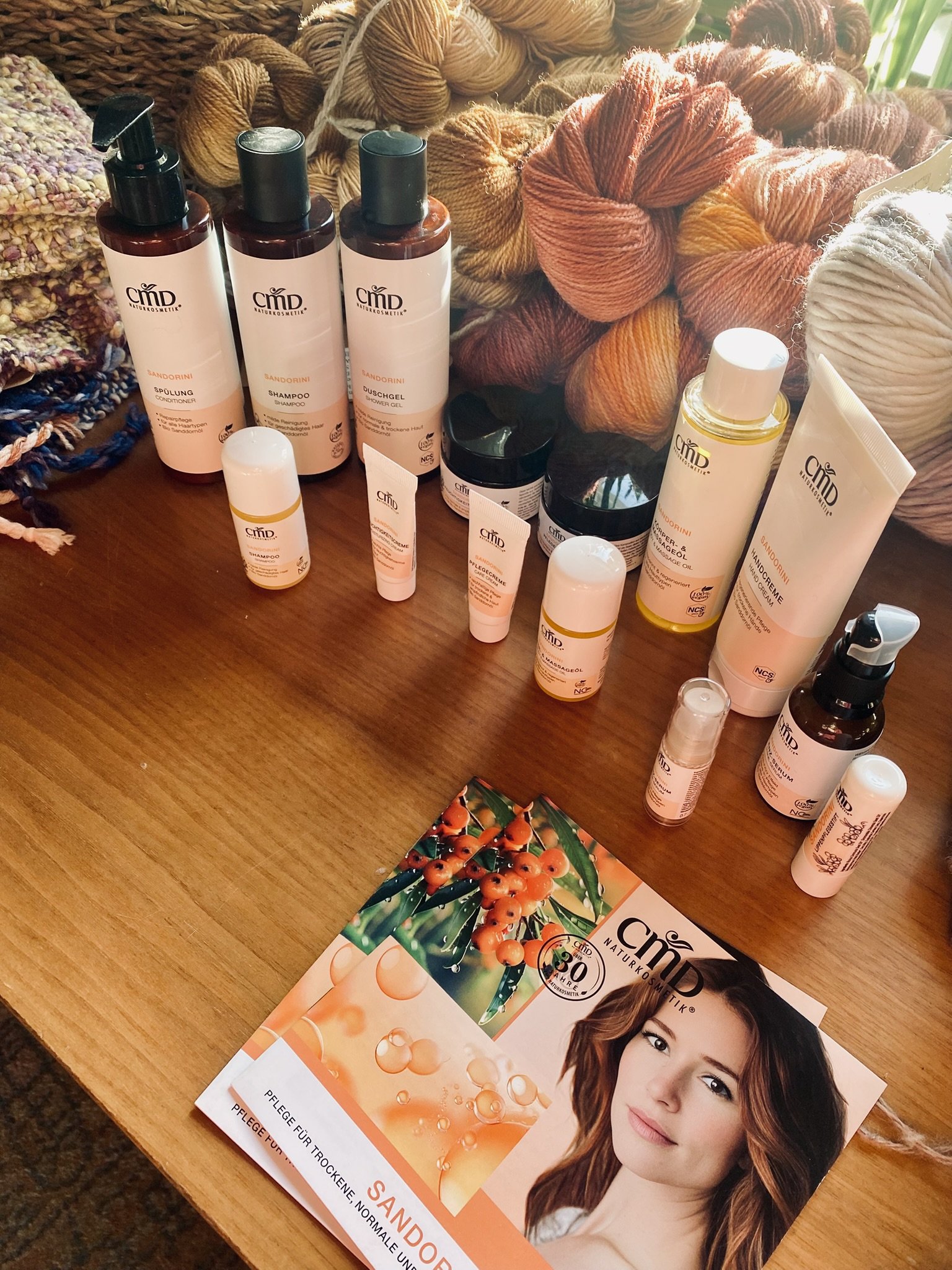 Verschiedene Pflegeprodukte wie Shampoo, Duschgel, Cremes, Öle und Lotionen liegen auf einem Holztisch. Im Hintergrund sind Handstrickgarne in verschiedenen Farben. Ein Flyer mit einer Frau und Beeren ist auf dem Tisch.