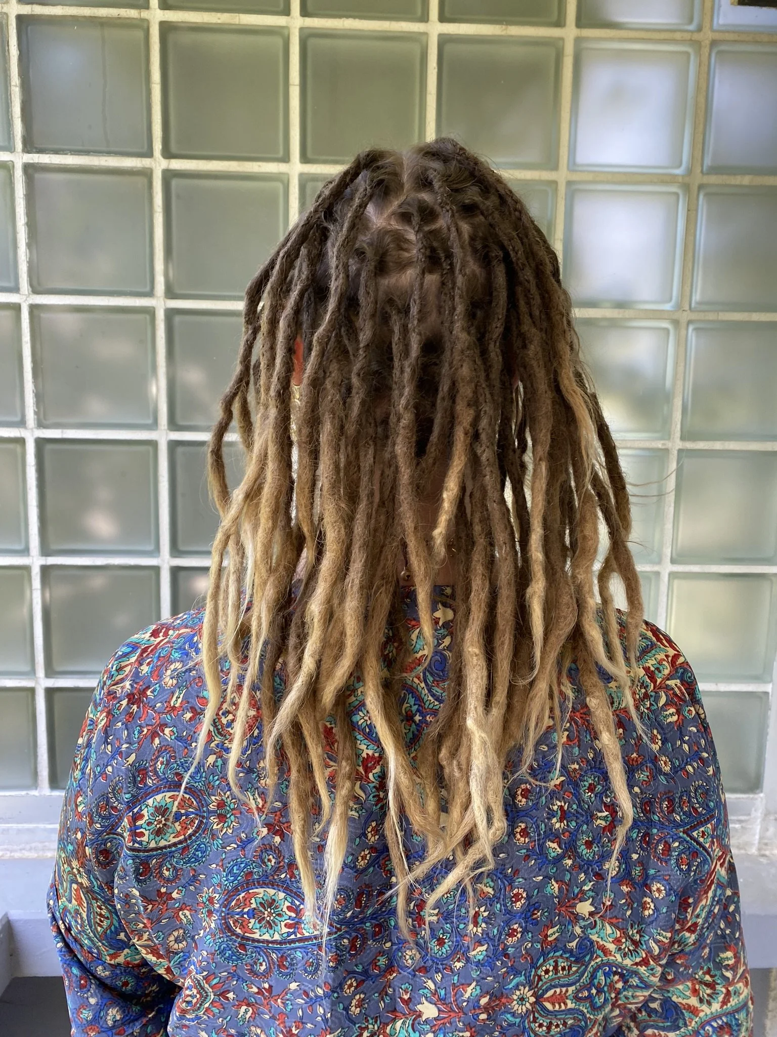 Eine Person mit langem, gewellten Dreadlocks trägt eine bunte, paisleyartige Jacke vor einem Hintergrund aus Glasblöcken.