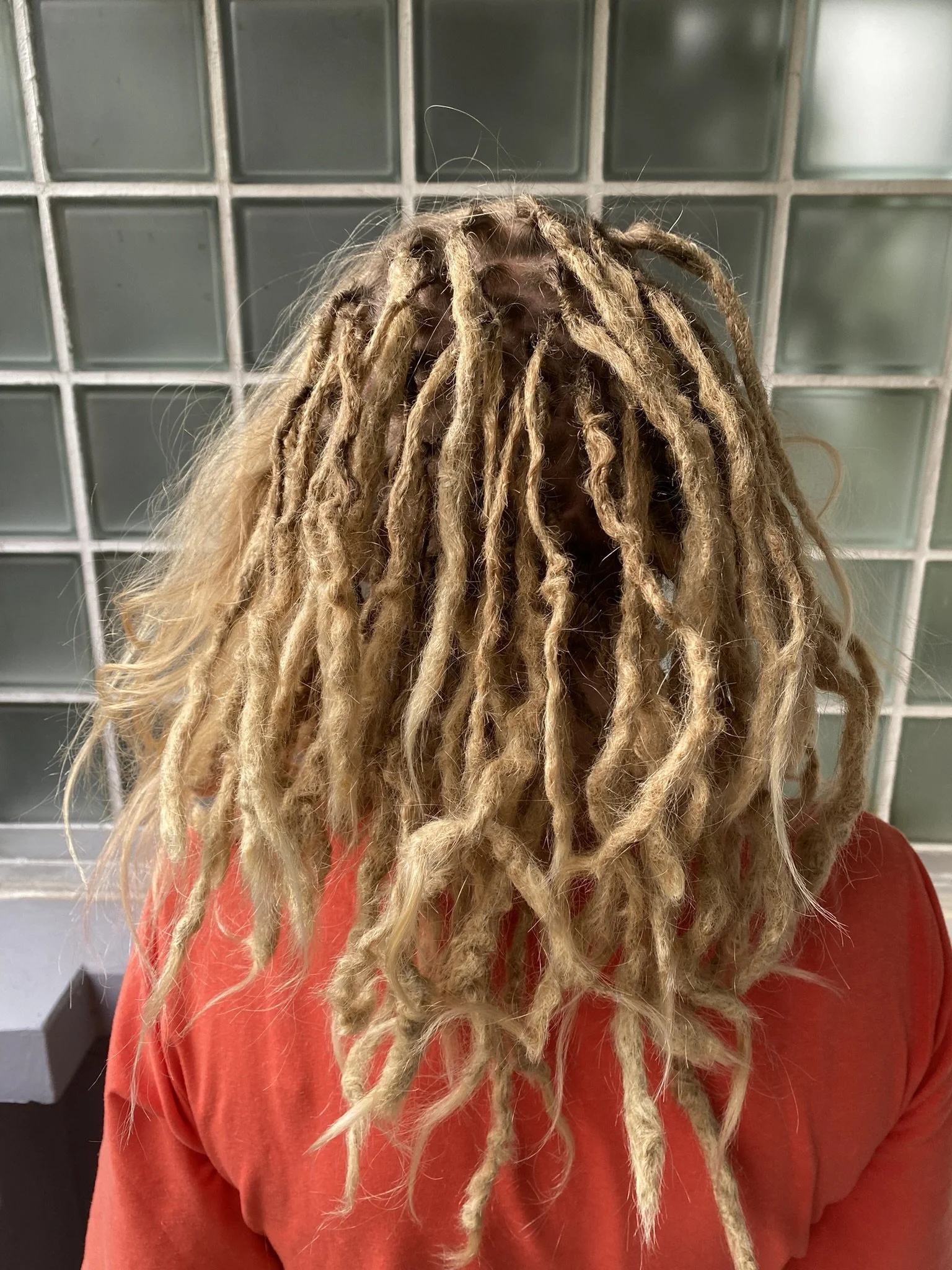 Person mit langem, blonden Dreadlocks, trägt eine orange Jacke, steht vor einer Wand aus Glasblöcken.