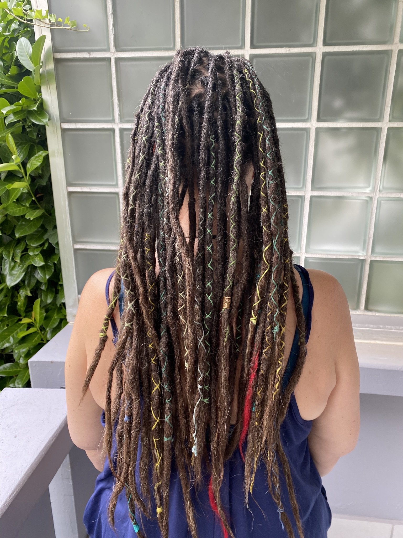 Eine Person mit langen Dreadlocks, die zum Rücken zeigt, vor einer Wand aus Glasblöcken und begrünten Flächen.