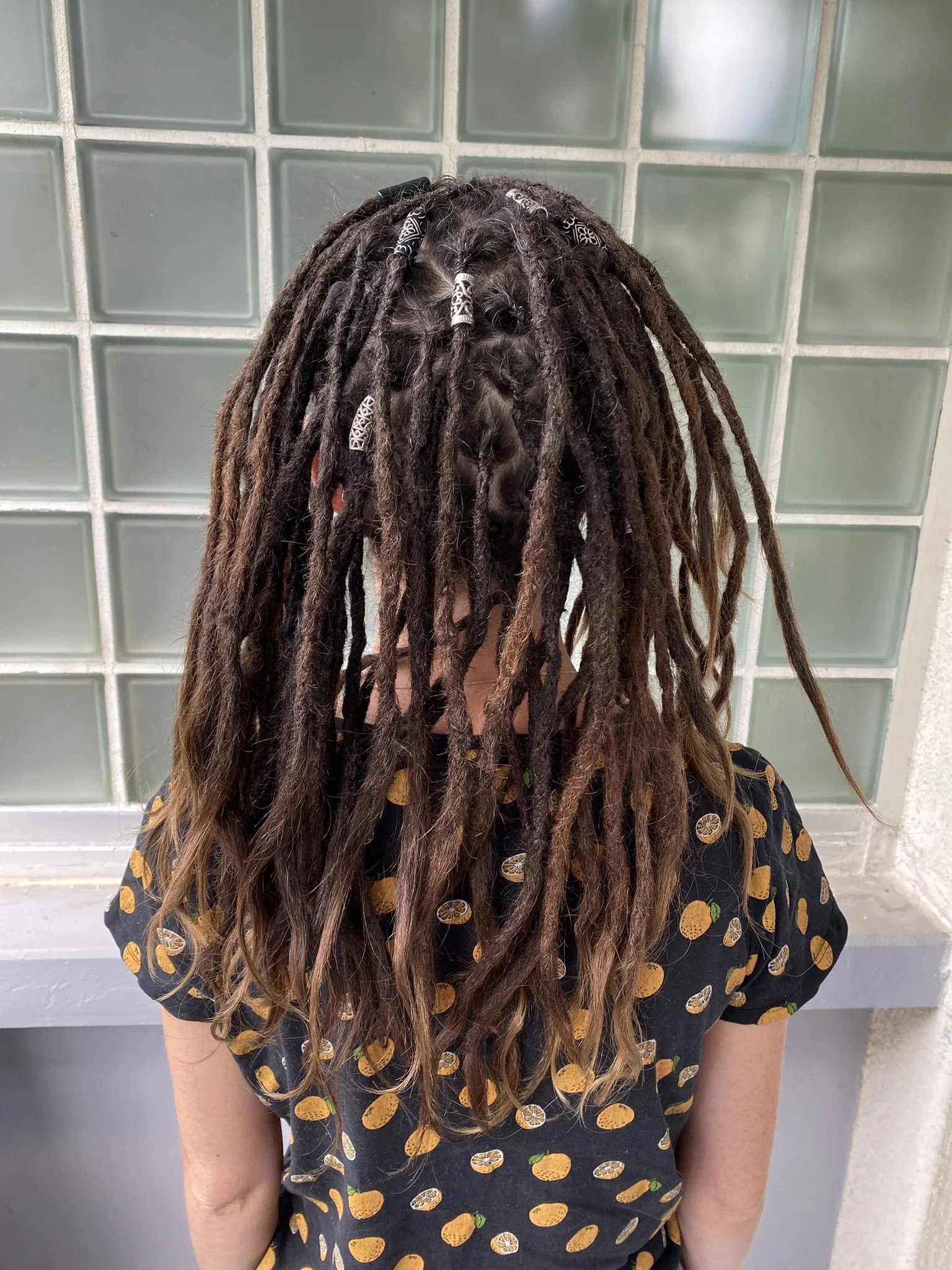 Person mit dreadlocks, teilweise mit schwarzen und weißen Haarbändern, steht vor einer Glassteinwand, trägt ein schwarzes Hemd mit Orangenscheibenmuster.