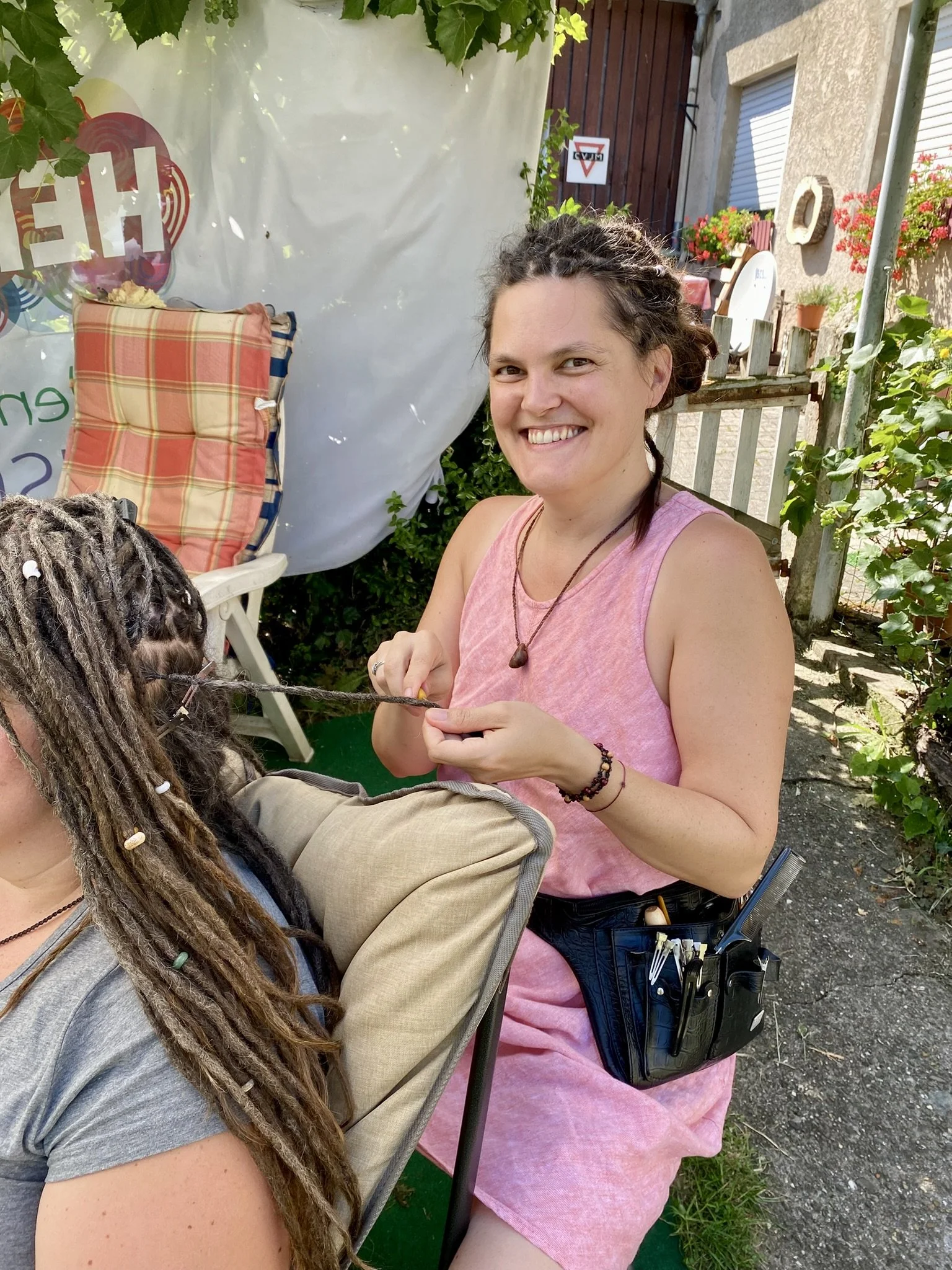 Eine lachende Frau mit lockigen braunen Haaren, im pinken Kleid, beim Flechten der Haare einer anderen Person mit dreadlocks, im Freien bei Sonnenschein.
