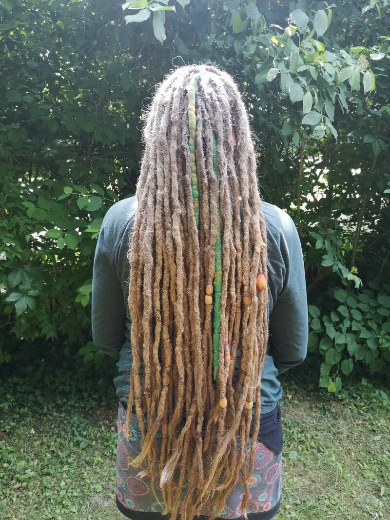 Person mit langen Dreadlocks vor einem grünen Busch.
