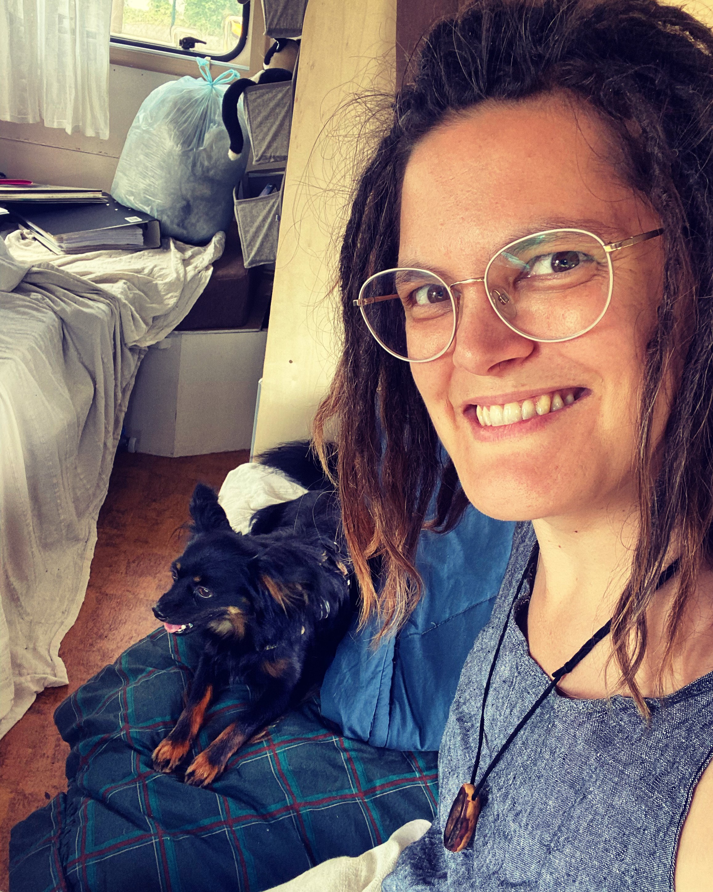 Eine Frau trägt eine Brille mit runden Gläsern und sitzt mit einem schwarzen, braunen Hund auf den Knien. Das Zimmer hat ein unordentliches Bett und ein Fenster im Hintergrund.