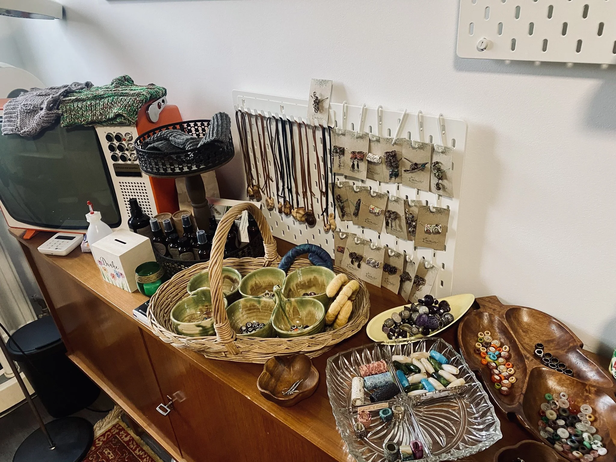 Ein Tisch mit Schmuck, Halsketten, Ringen, Armbändern und Brillen. Es sind verschiedene Schalen und Behälter mit Perlen, Steinen und anderen Schmuckmaterialien. An der Wand eine Schmuckhalter mit Anhängern und Ohrringen.