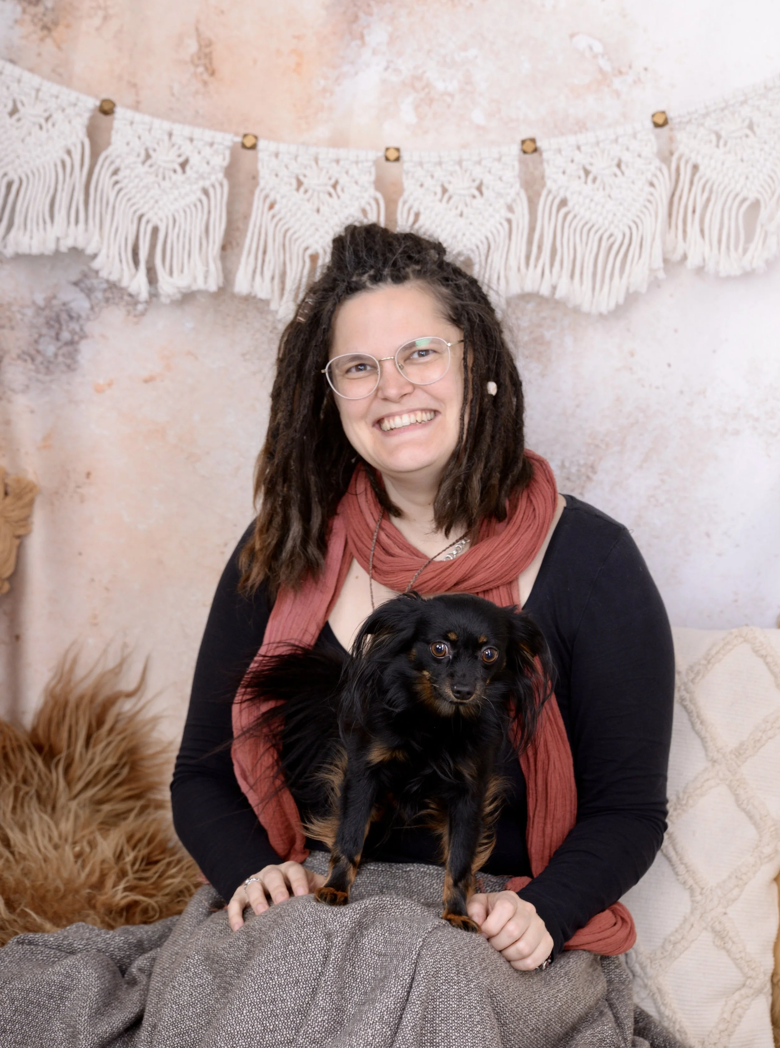 Eine lachende Frau mit Brille und roten Schal sitzt auf einem Sofa, hält eine kleine schwarze und braune Hund. Hinter ihnen hängen dekorative Wand-Textilien.