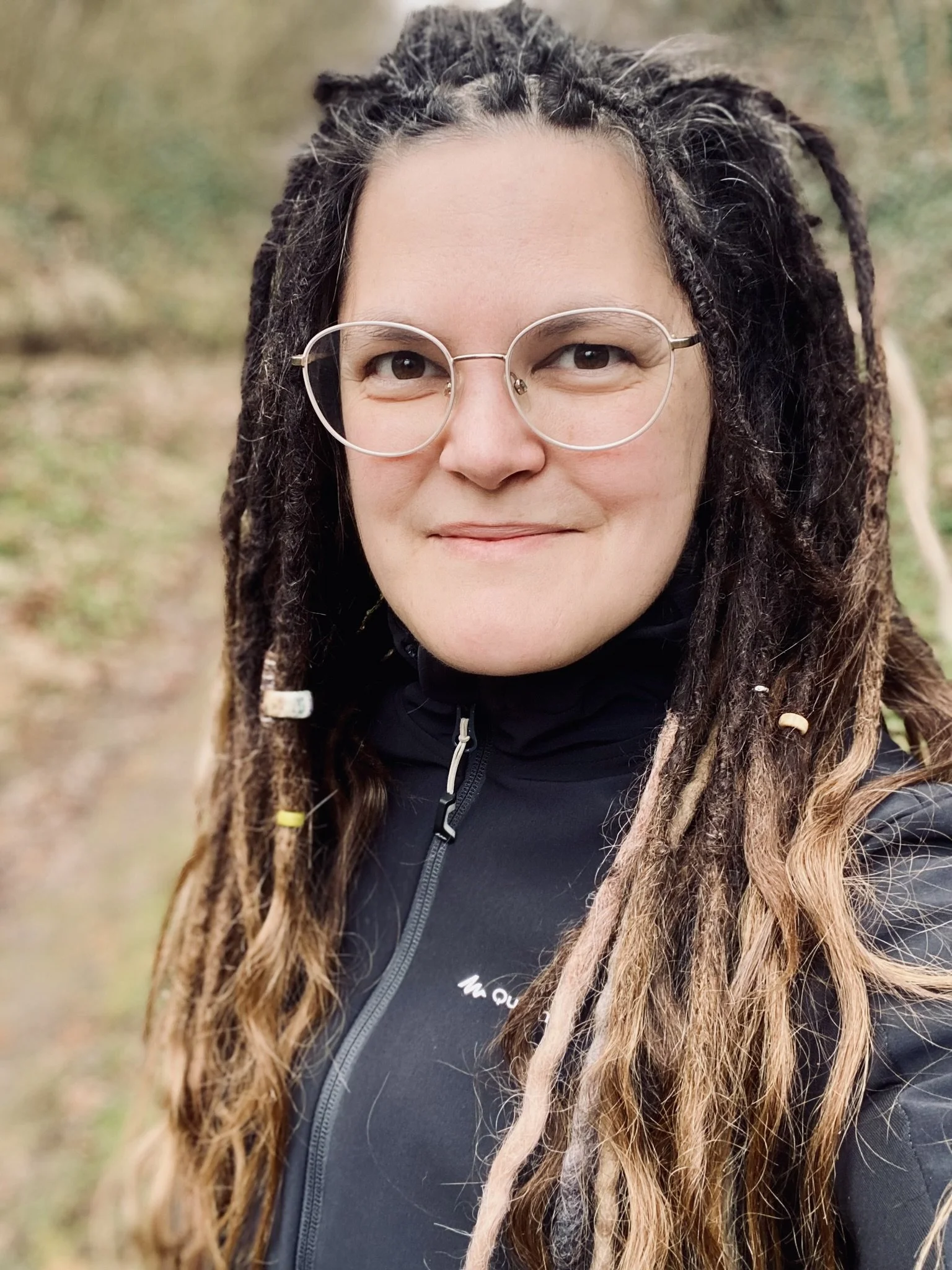 Eine Frau mit dreadlocks und Brille, trägt eine schwarze Jacke und steht im Freien.