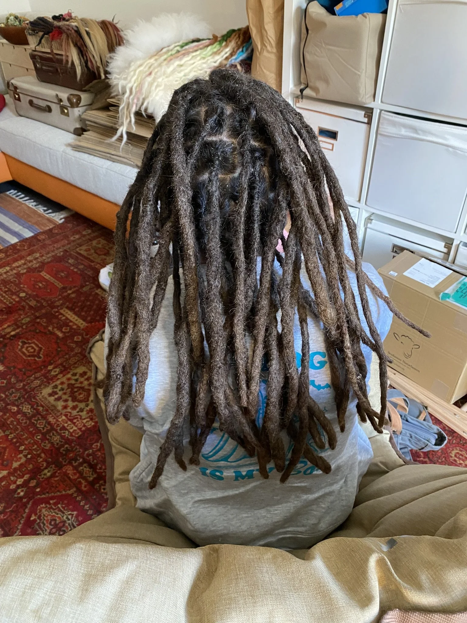 Person mit langen dreadlocks sitzend in einem Wohnzimmer, im Hintergrund Couch, Koffer, Decken und Kartons.
