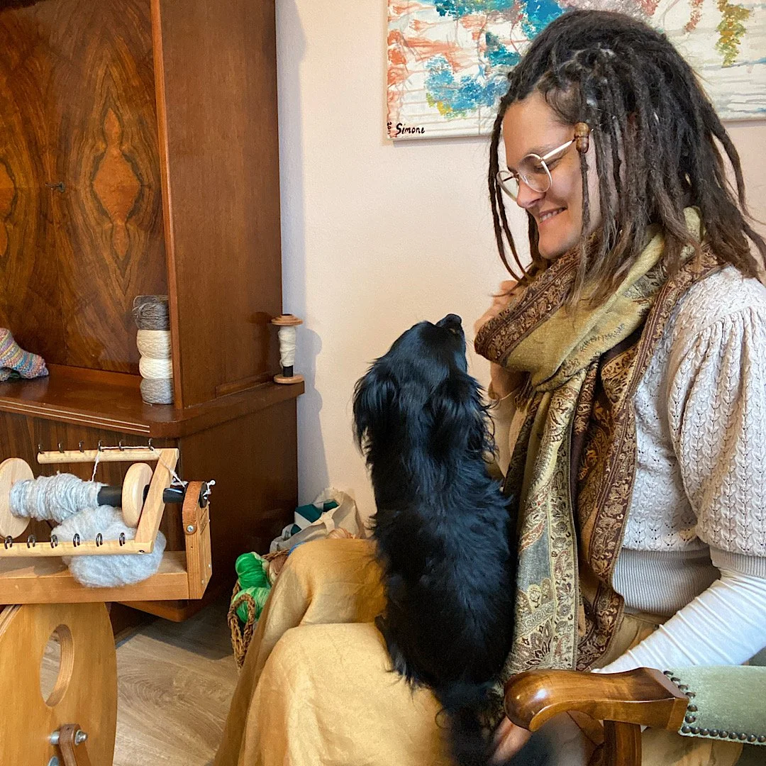 Frau mit Brille und lockigen Haaren sitzt auf einem Stuhl, hält einen schwarzen Hund auf ihrem Schoß, beide schauen sich an und lächeln.