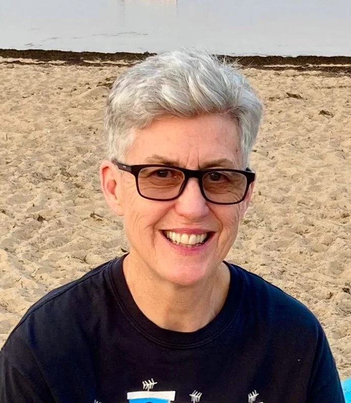 A picture of Sandy Bailey with short gray hair and glasses sitting on a sandy beach with water in the background, near Boston Massachusetts.