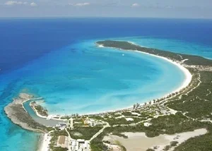 A crescent-shaped tropical beach with clear turquoise water, surrounding a lagoon, with green land and buildings nearby.