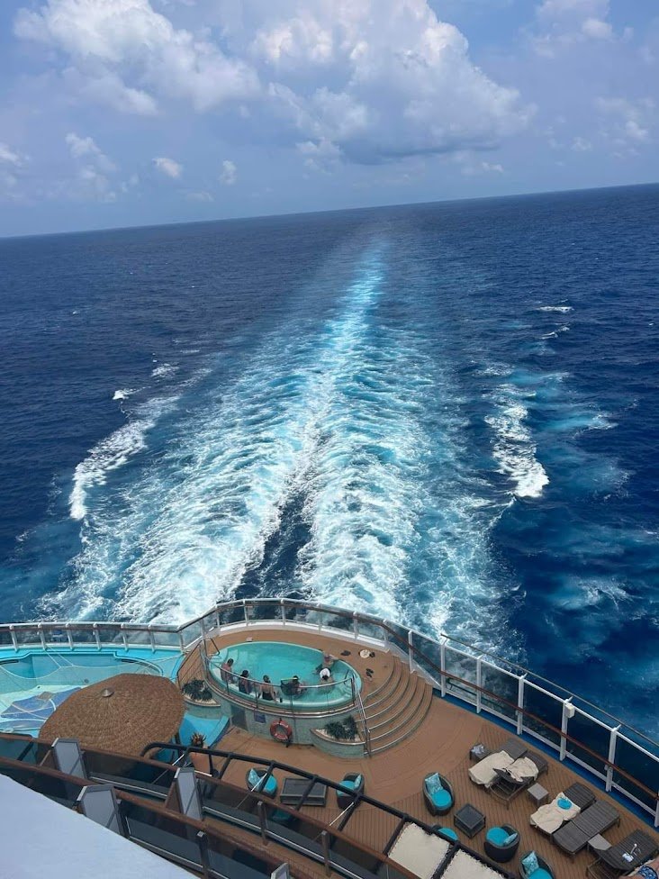 View from a cruise ship showing the ocean and the ship's deck with a hot tub, lounge chairs, and a wake trail in the water behind the ship.