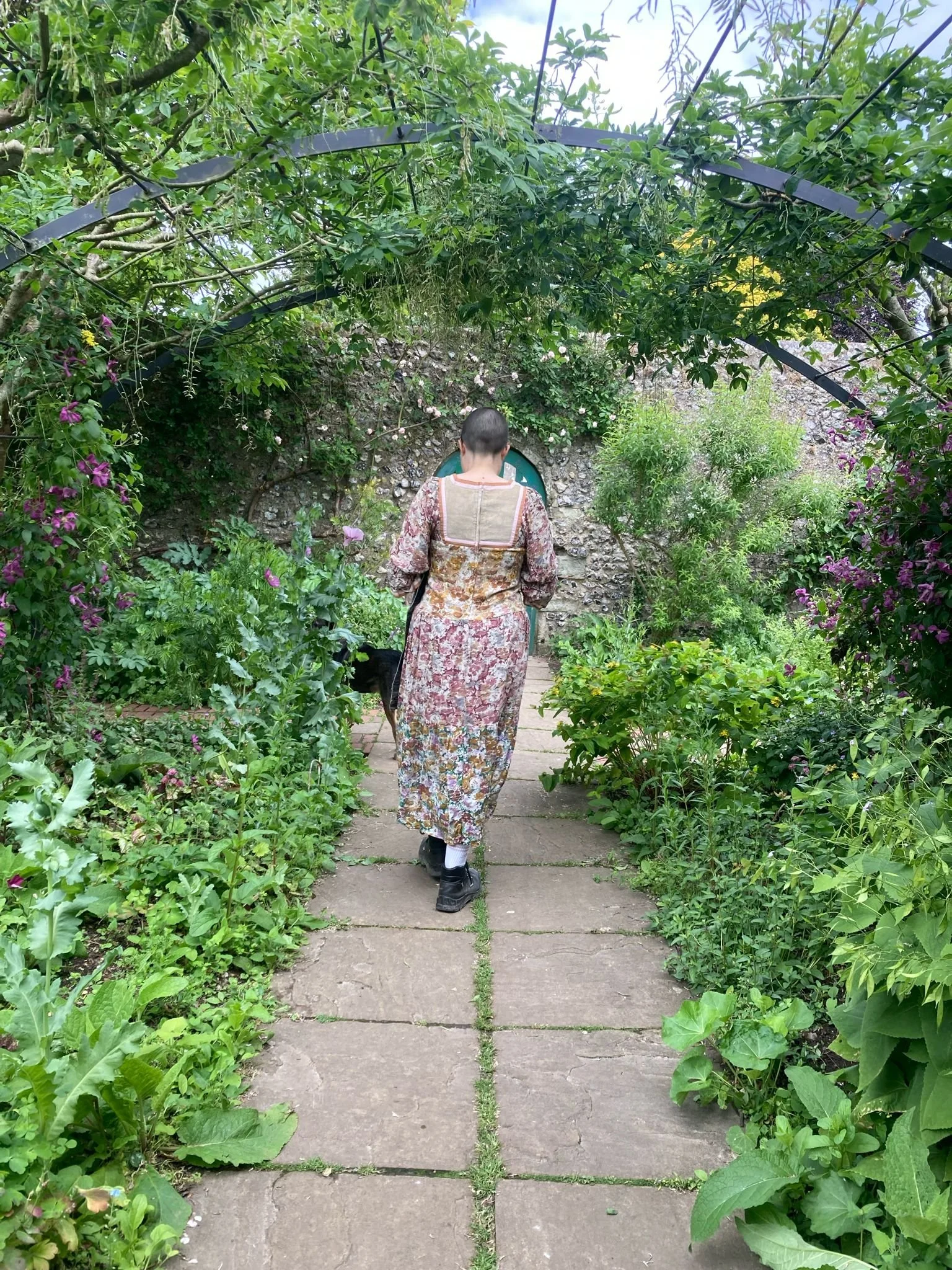 A person walking on a pathway surrounded by lush green plants and flowers in a garden.