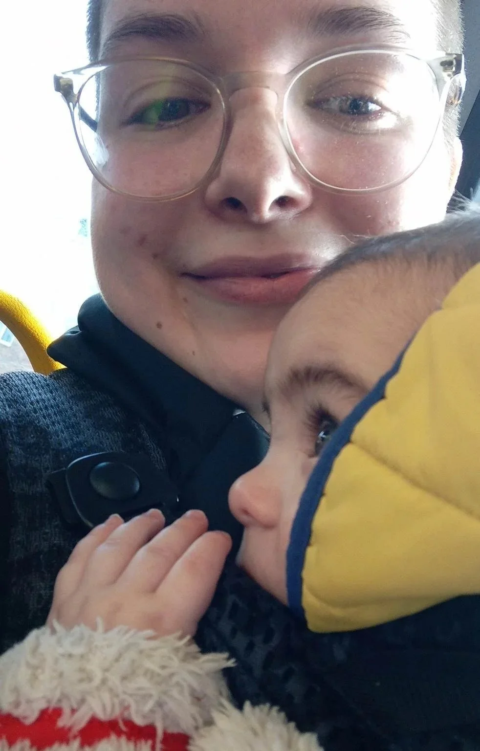 Close-up of a person with glasses and a slight smile holding a young child with a yellow jacket, both in a close embrace.