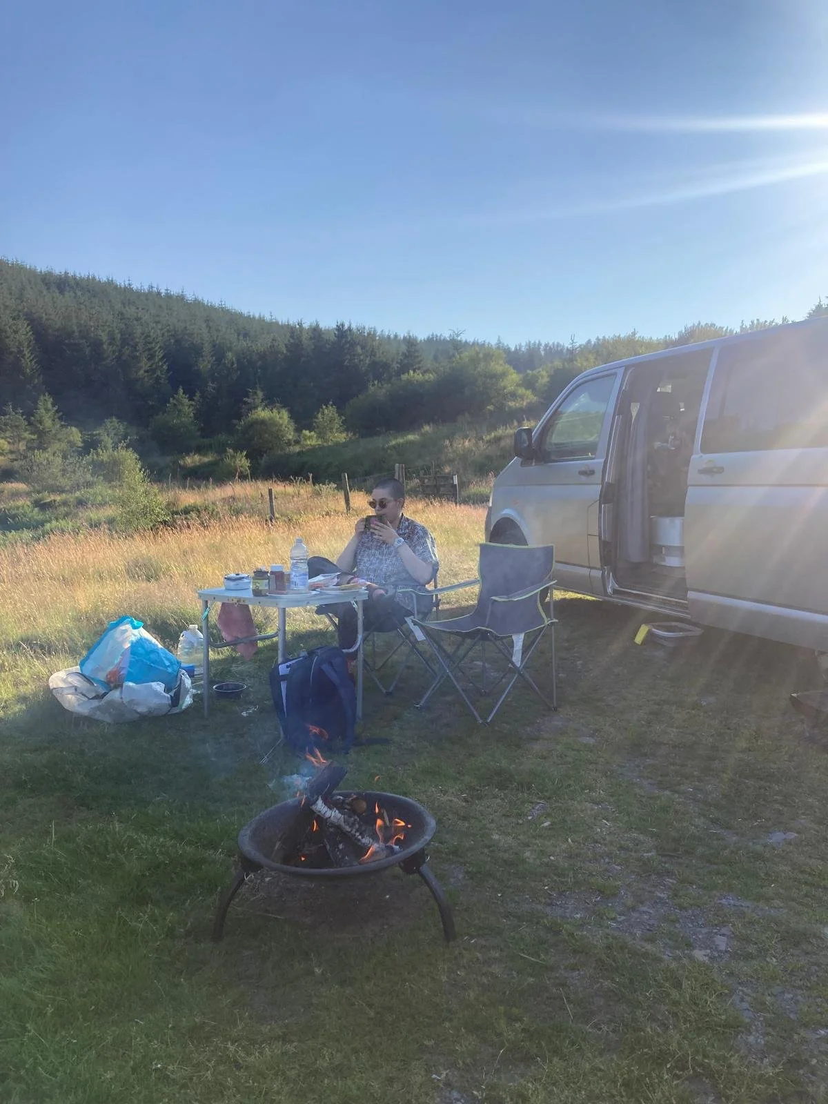 A person sitting at a camping table outdoors near a van, with camping gear and an open fire pit in a grassy field with trees and hills in the background on a sunny day.