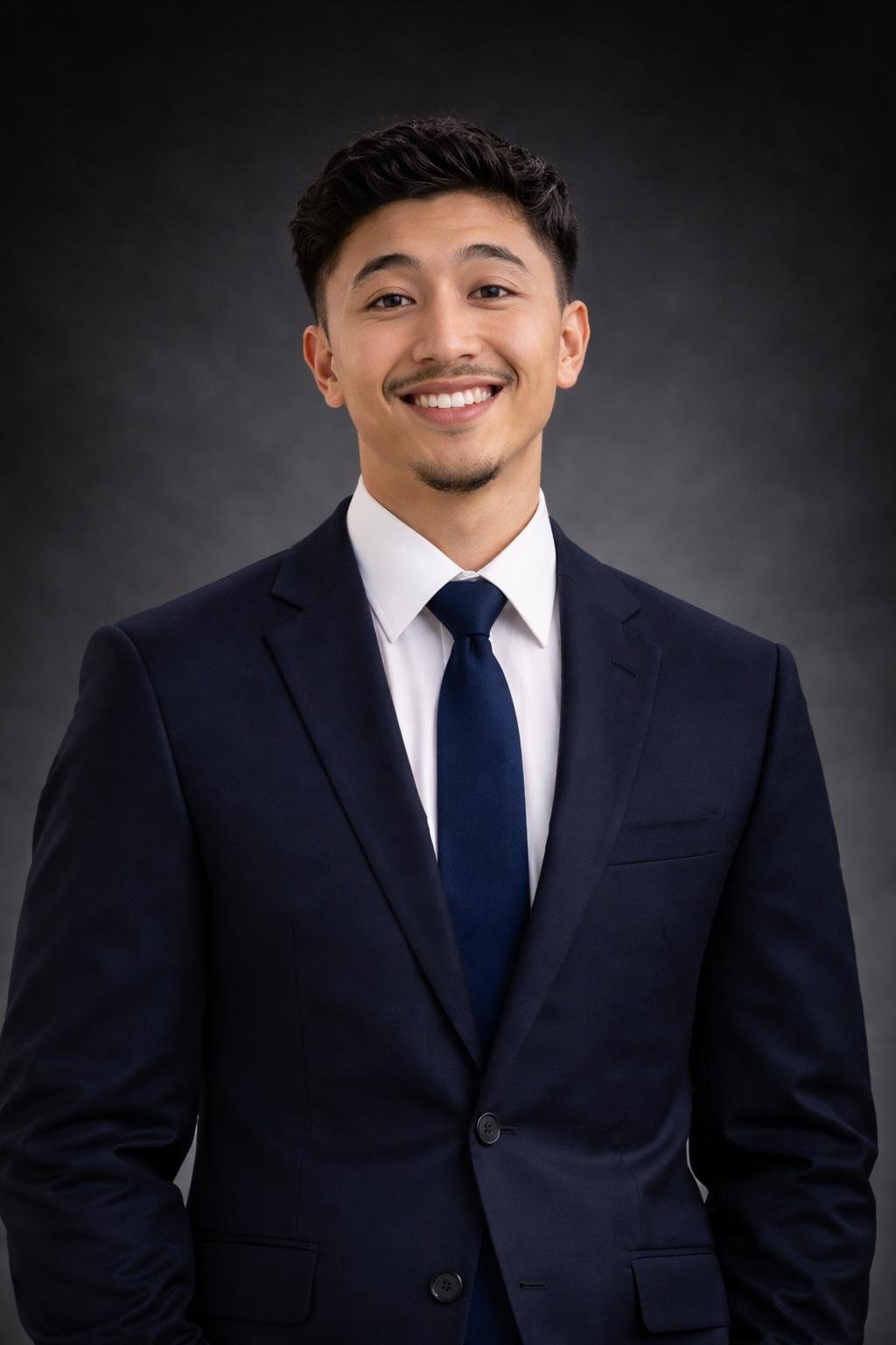 A young man with short dark hair, wearing a navy blue suit, white shirt, and navy tie, smiling against a dark grey background.