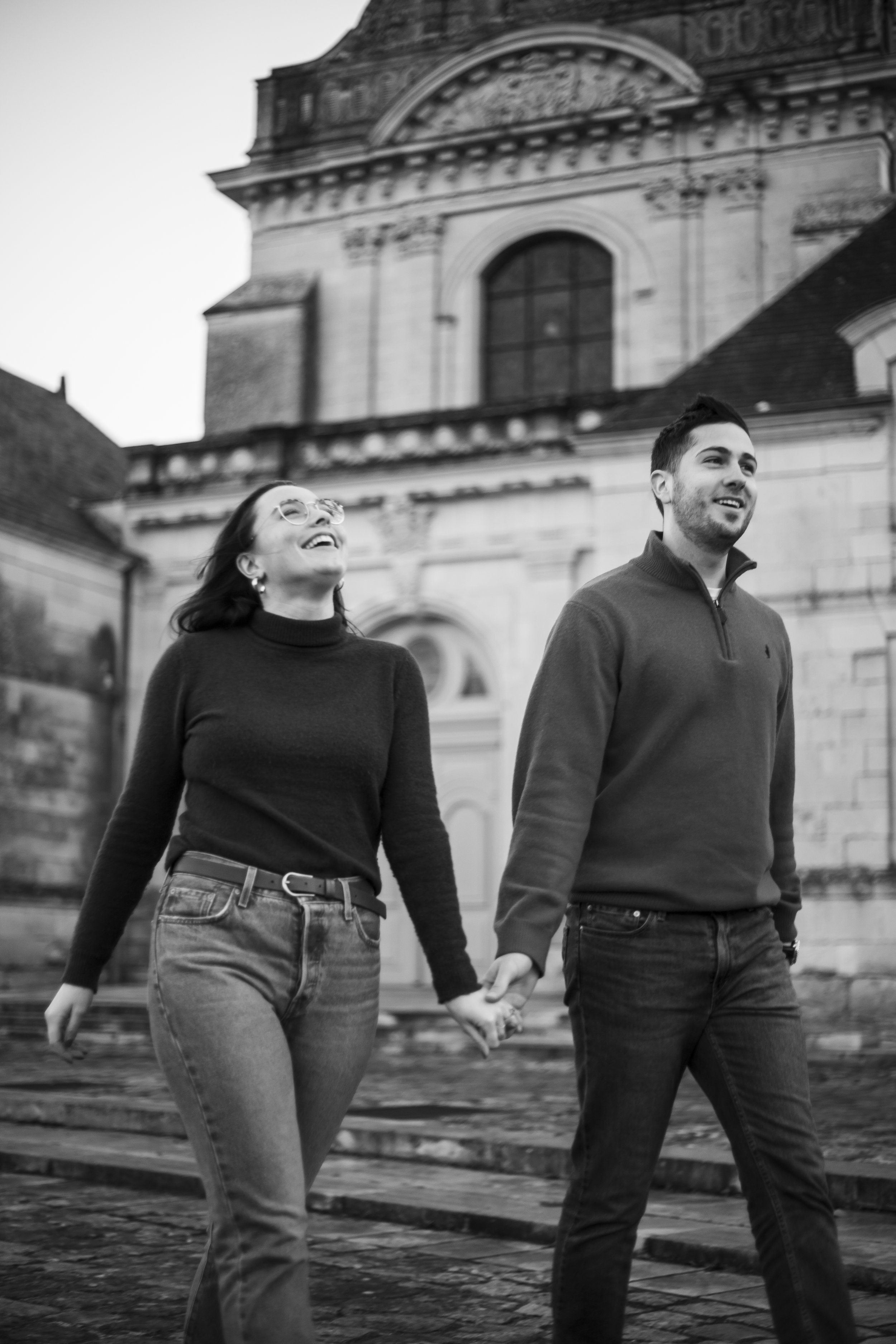 Un couple marche main dans la main devant une vieille église en pierre, souriant et détendu, en noir et blanc.