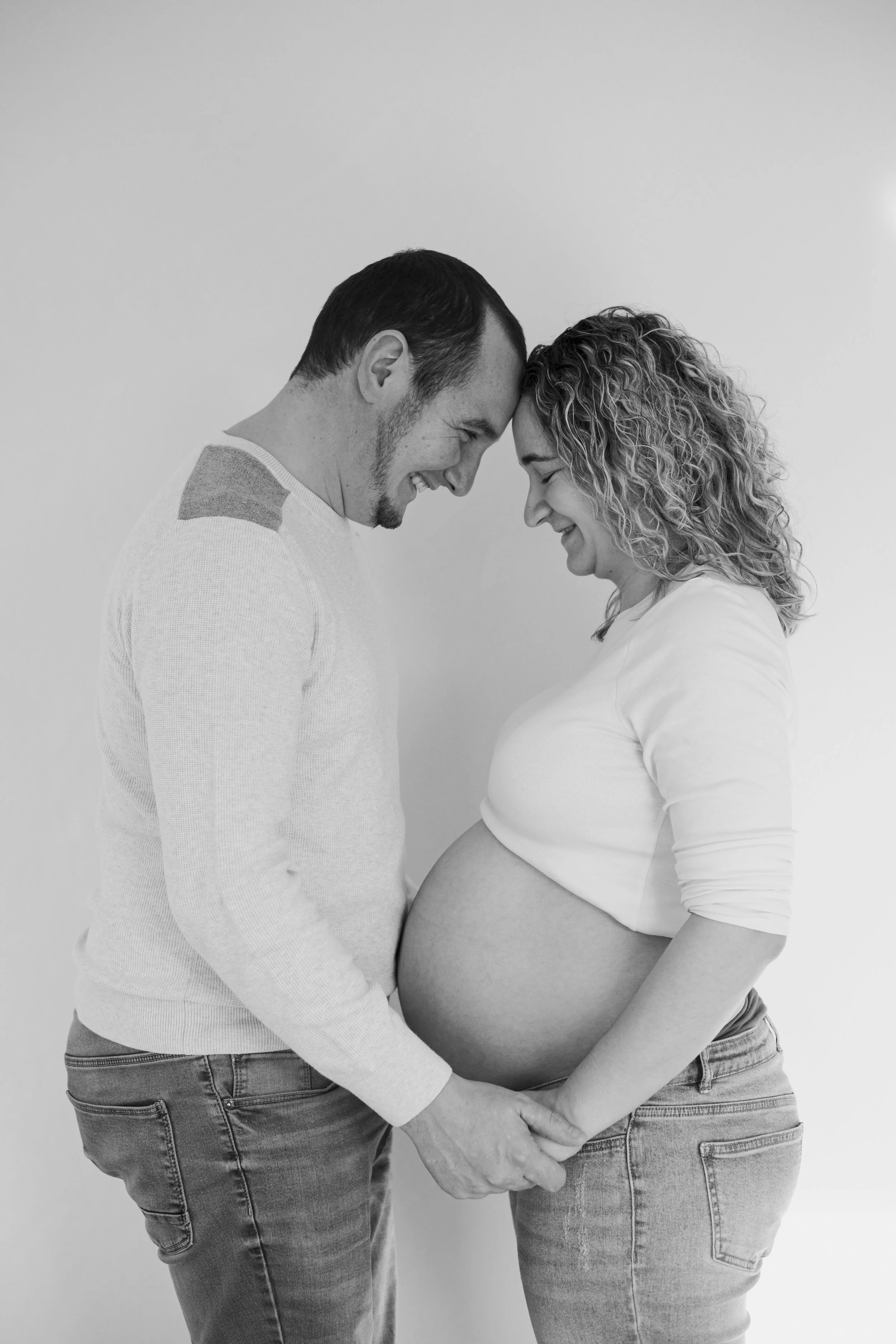 Un couple est debout face à face, les fronts touchant, la femme est enceinte, ils se tiennent par les mains, souriant avec tendresse dans une photo en noir et blanc.