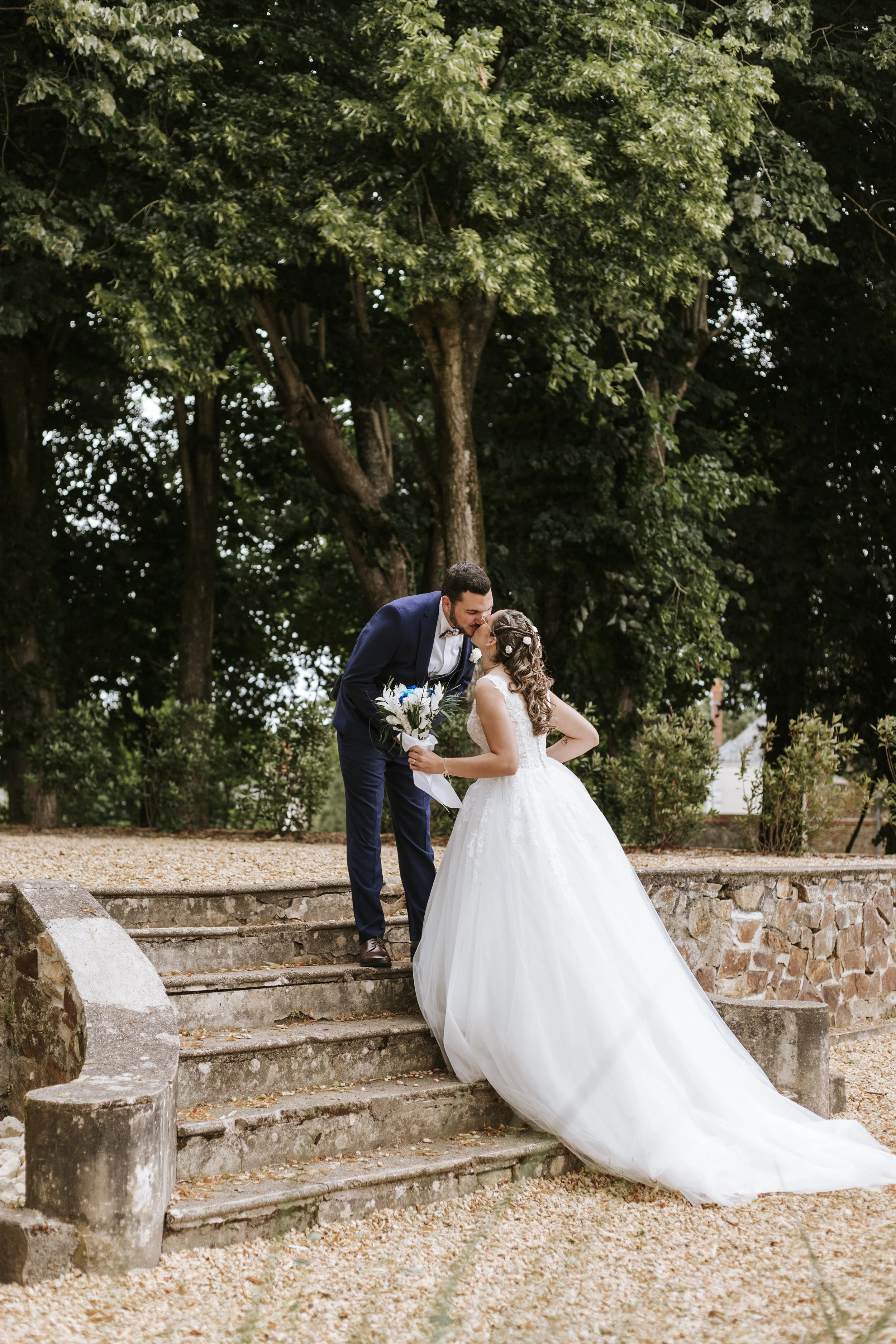 Un couple de mariés s'embrassant sur des marches en pierre dans un parc, avec de grands arbres en arrière-plan.