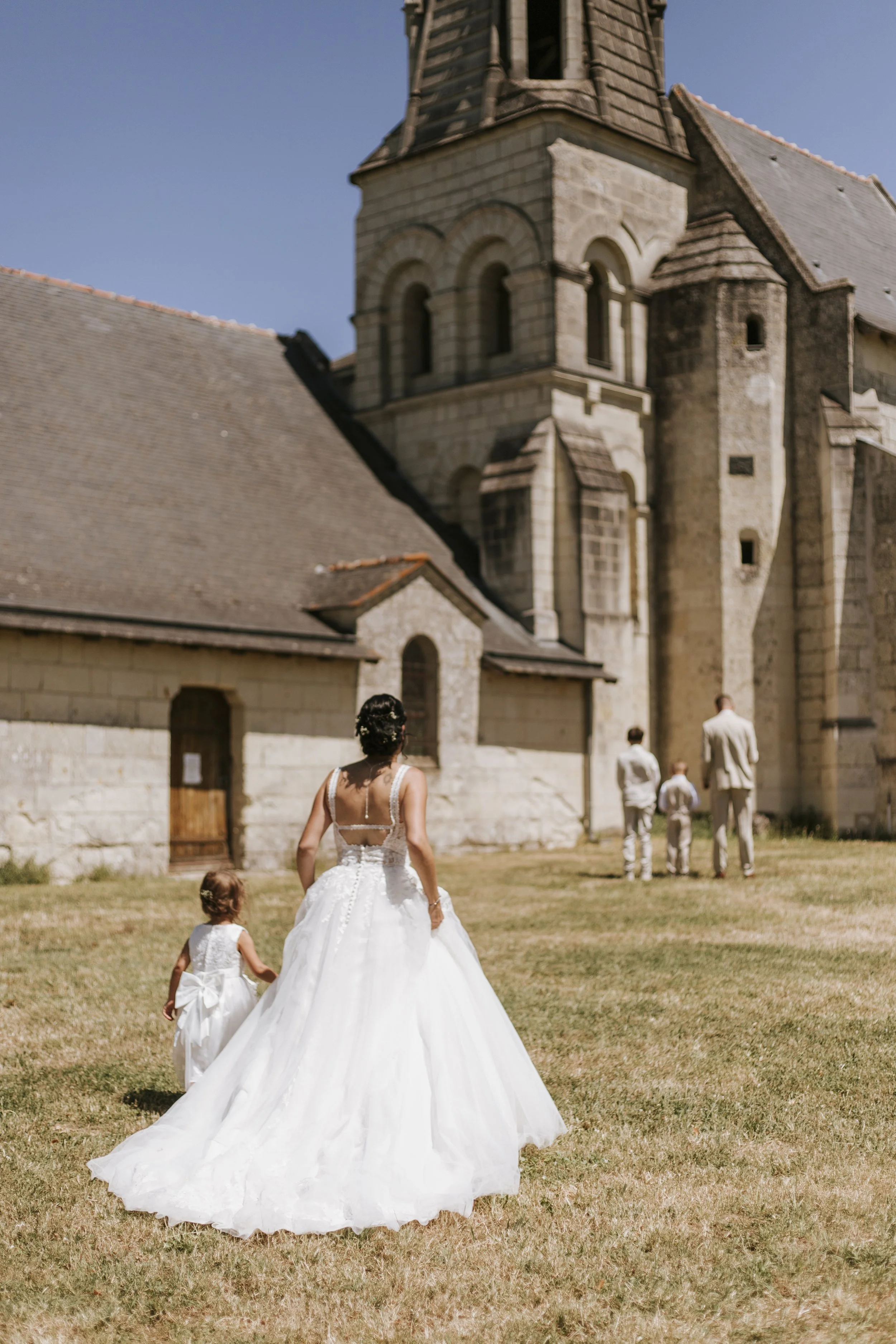 Une femme en robe de mariée, accompagnée de deux jeunes filles, marche vers une vieille église en pierre par une journée ensoleillée.