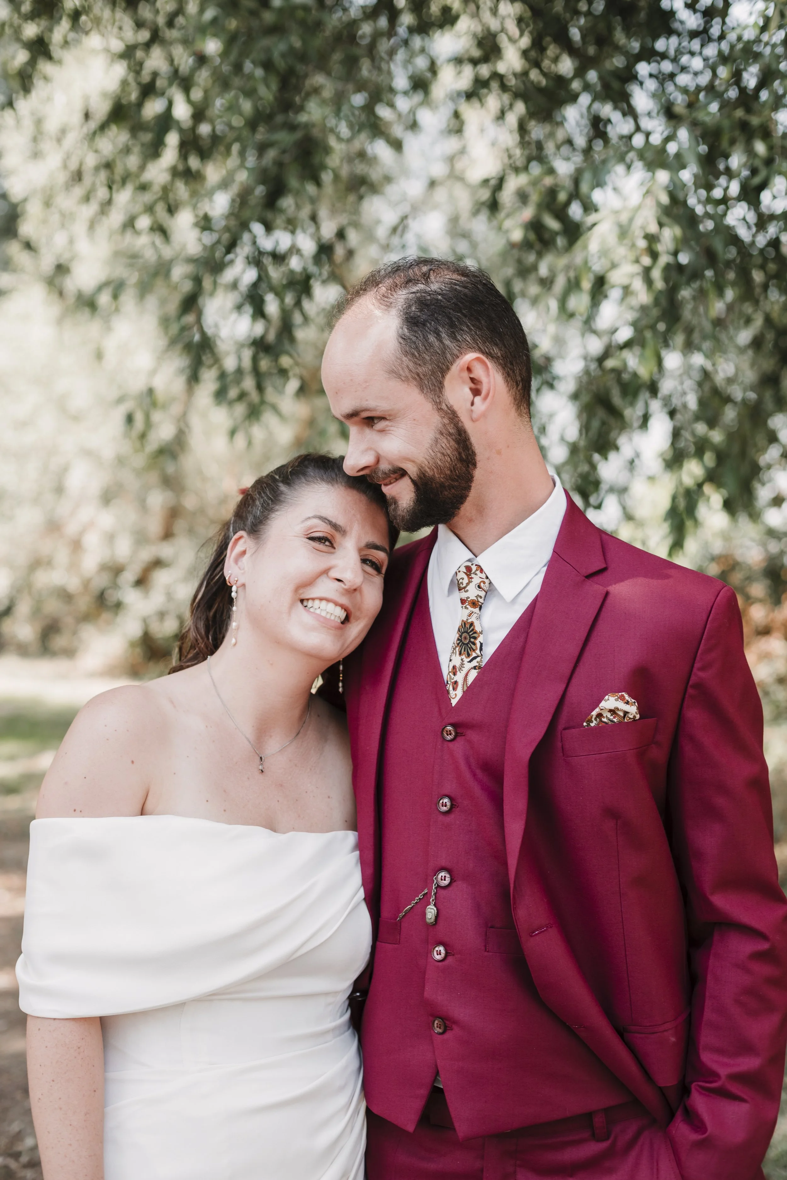 Un couple de mariage souriant, la femme en robe blanche et le homme en costume rouge, sous un arbre dans un cadre naturel.