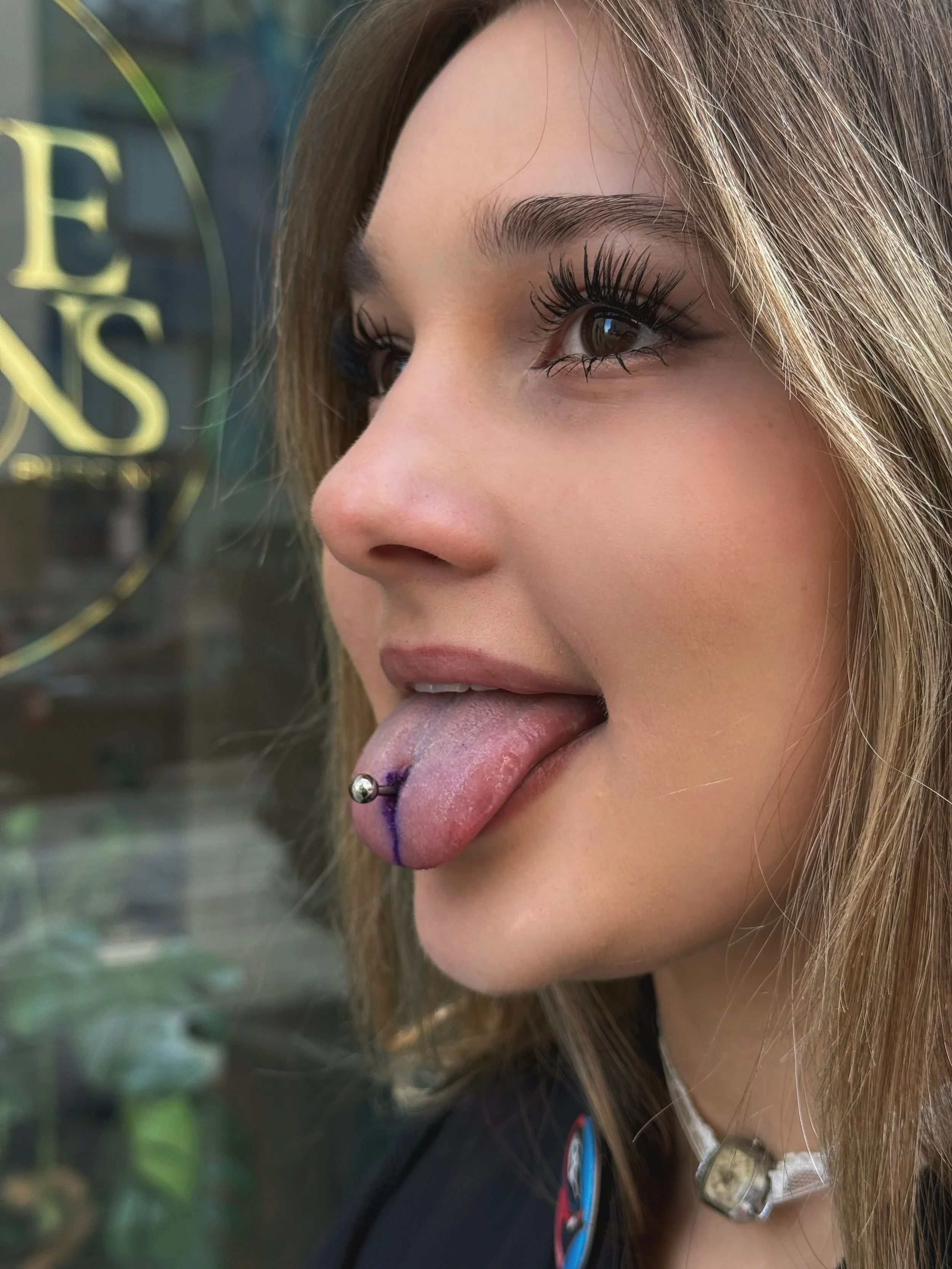 A young woman with brown hair, wearing makeup, has her tongue sticking out, showing a tongue piercing with a silver ball, and some purple and blue ink on her tongue, outside near a window.
