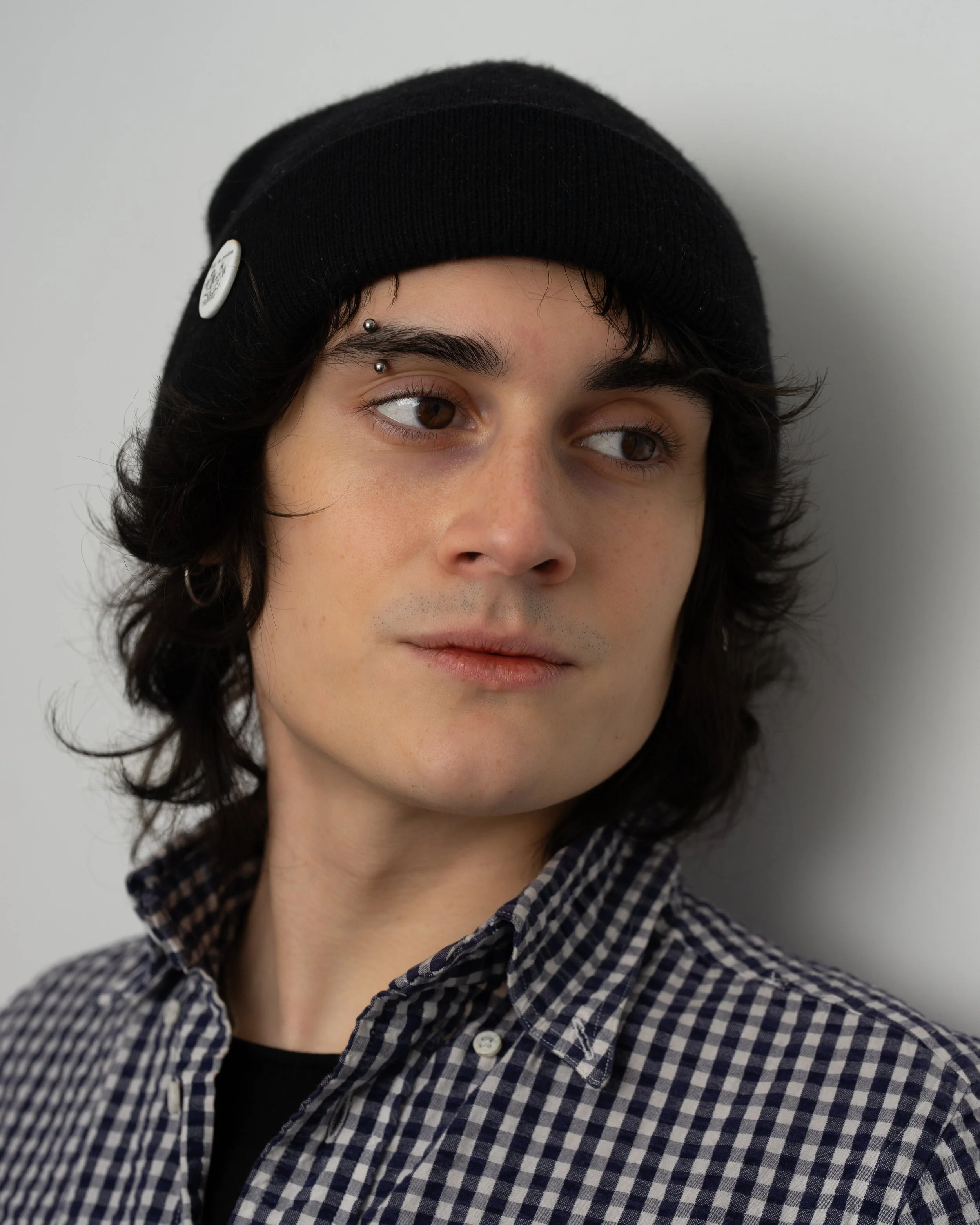 Close-up portrait of a young man with dark, wavy hair, wearing a black beanie and checkered shirt, looking to the right.