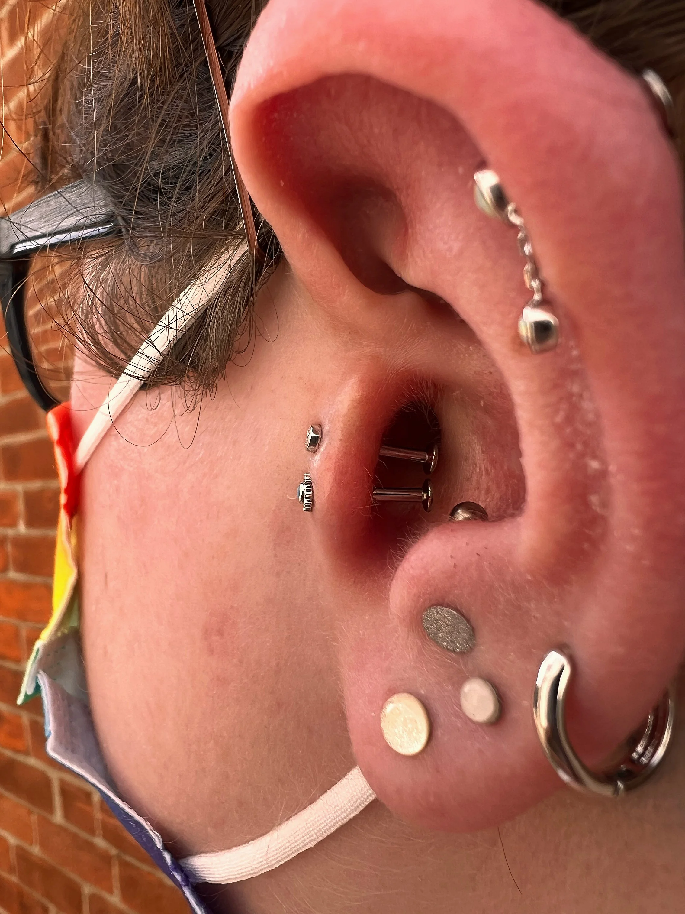 Close-up of a person's ear with multiple piercings and jewelry, including studs, hoops, and a chain, against a brick wall background.