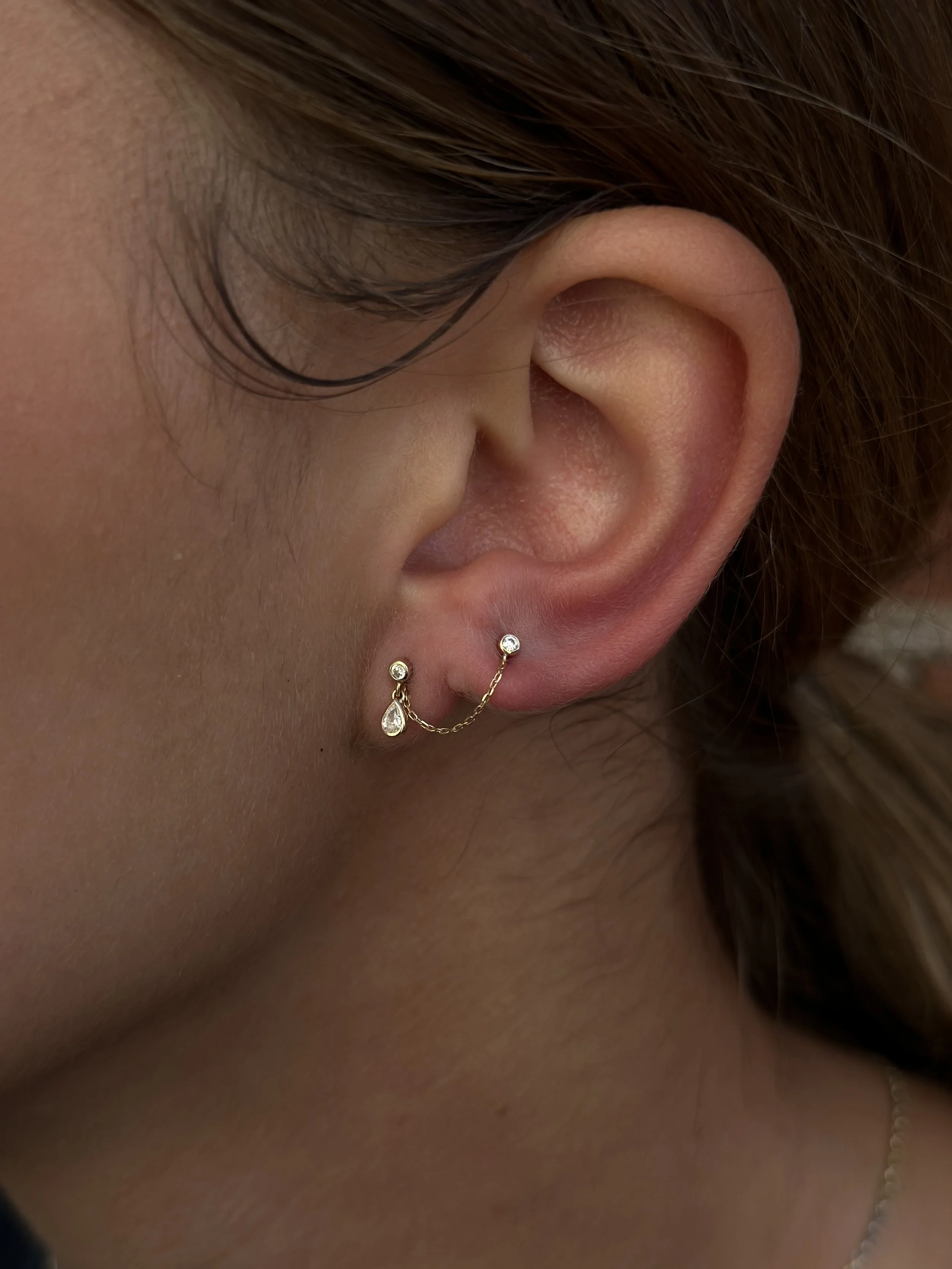 A close-up of a woman's ear with multiple earrings, including a gold chain dangle earring with a small diamond and a pearl-shaped charm.