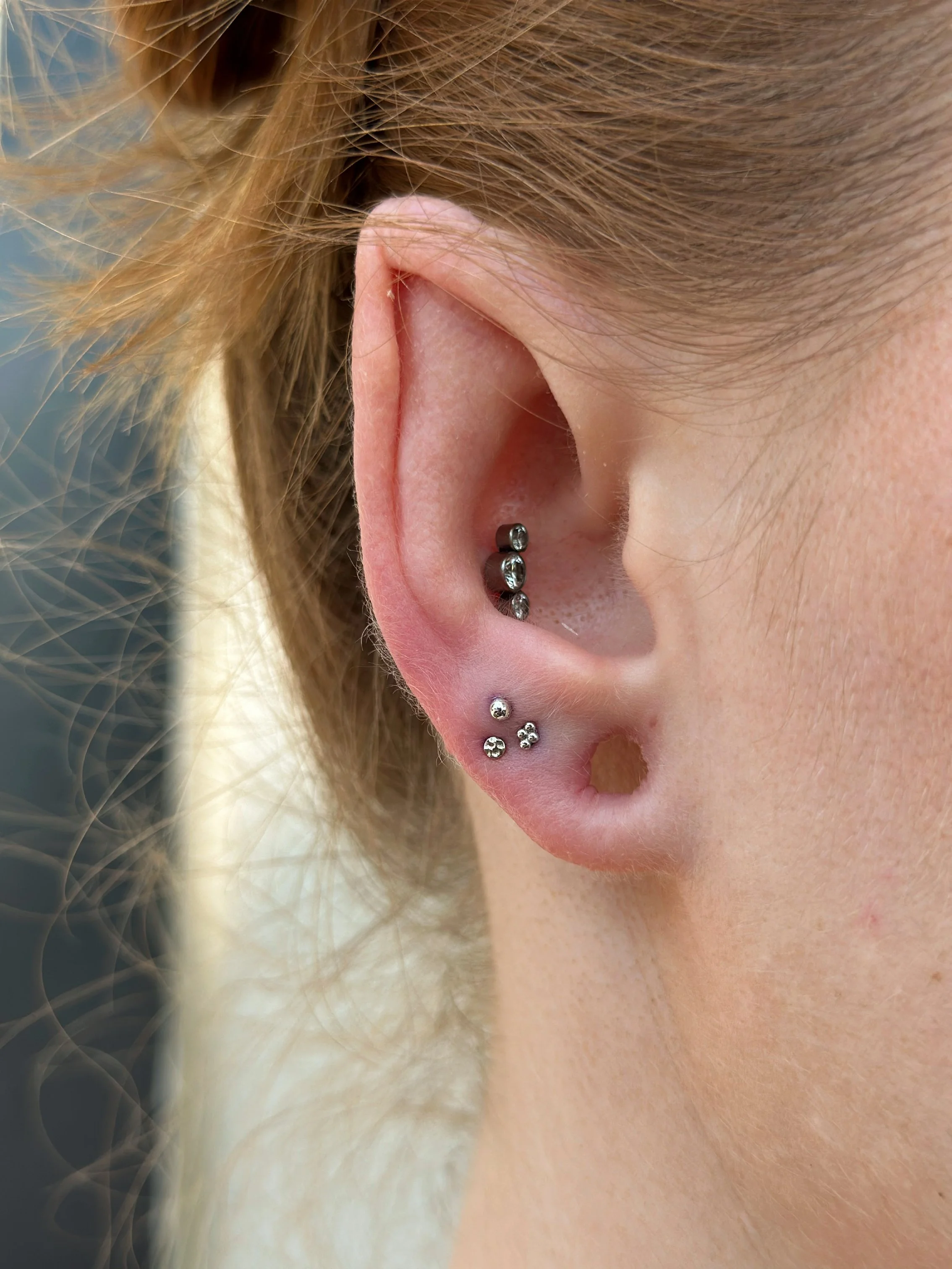 Close-up of a woman's ear with multiple piercings and jewelry, including a helix piercing with a dark gemstone and three lobe piercings with small studs.