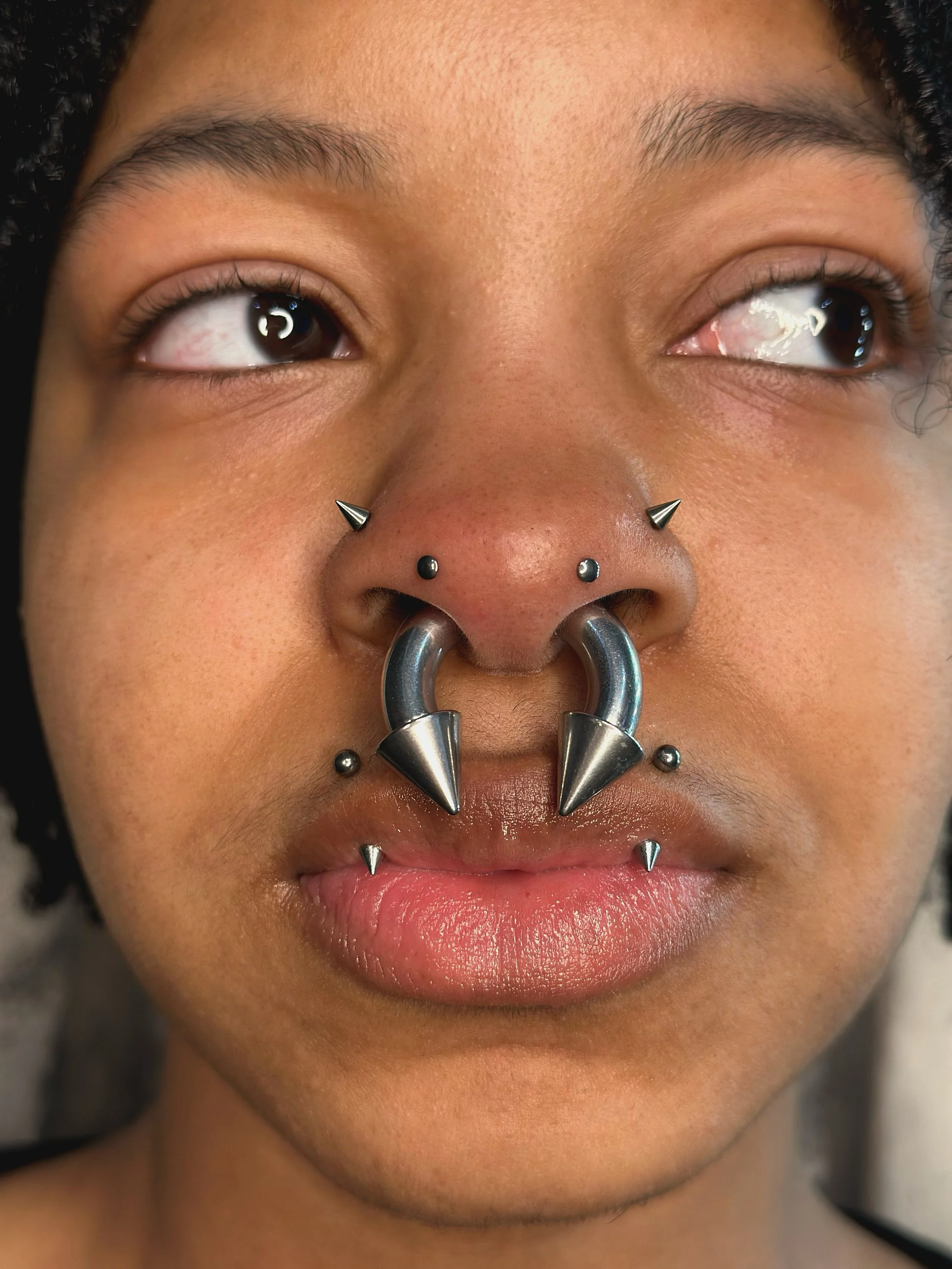 Close-up of a person with multiple facial piercings including a septum ring, nostril studs, eyebrow studs, and lip piercings, with dark curly hair.
