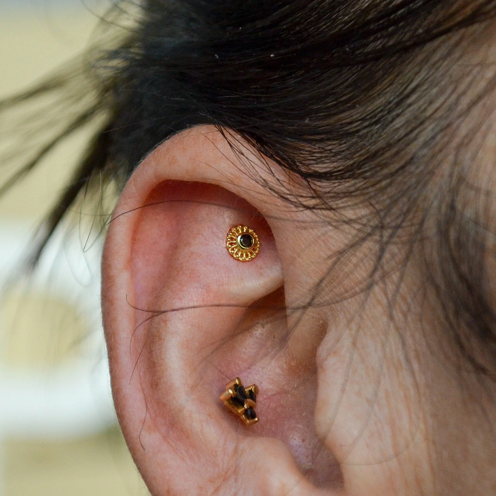 A close-up of a person's ear with a gold circular earring in the upper part and a gold hoop earring with black stones in the lower part of the ear.