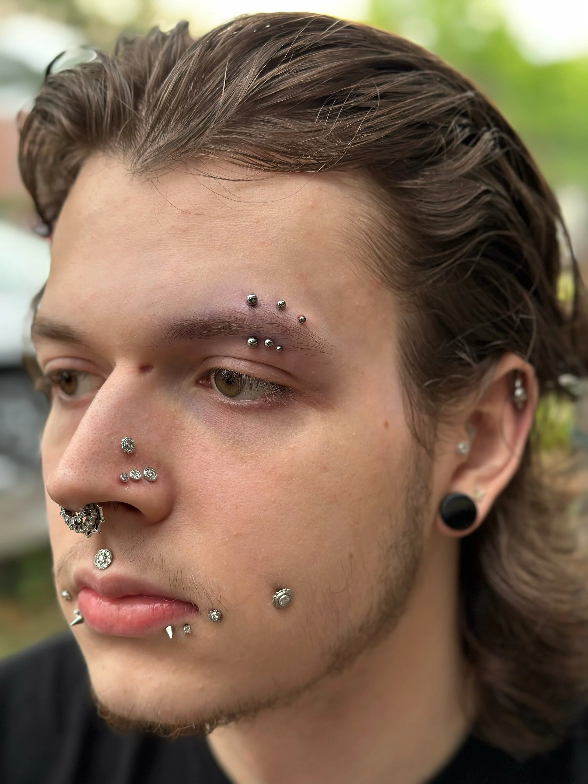 Close-up of a young man with multiple facial piercings, including piercings on eyebrows, nose, lips, and ears. He has brown hair and light skin.