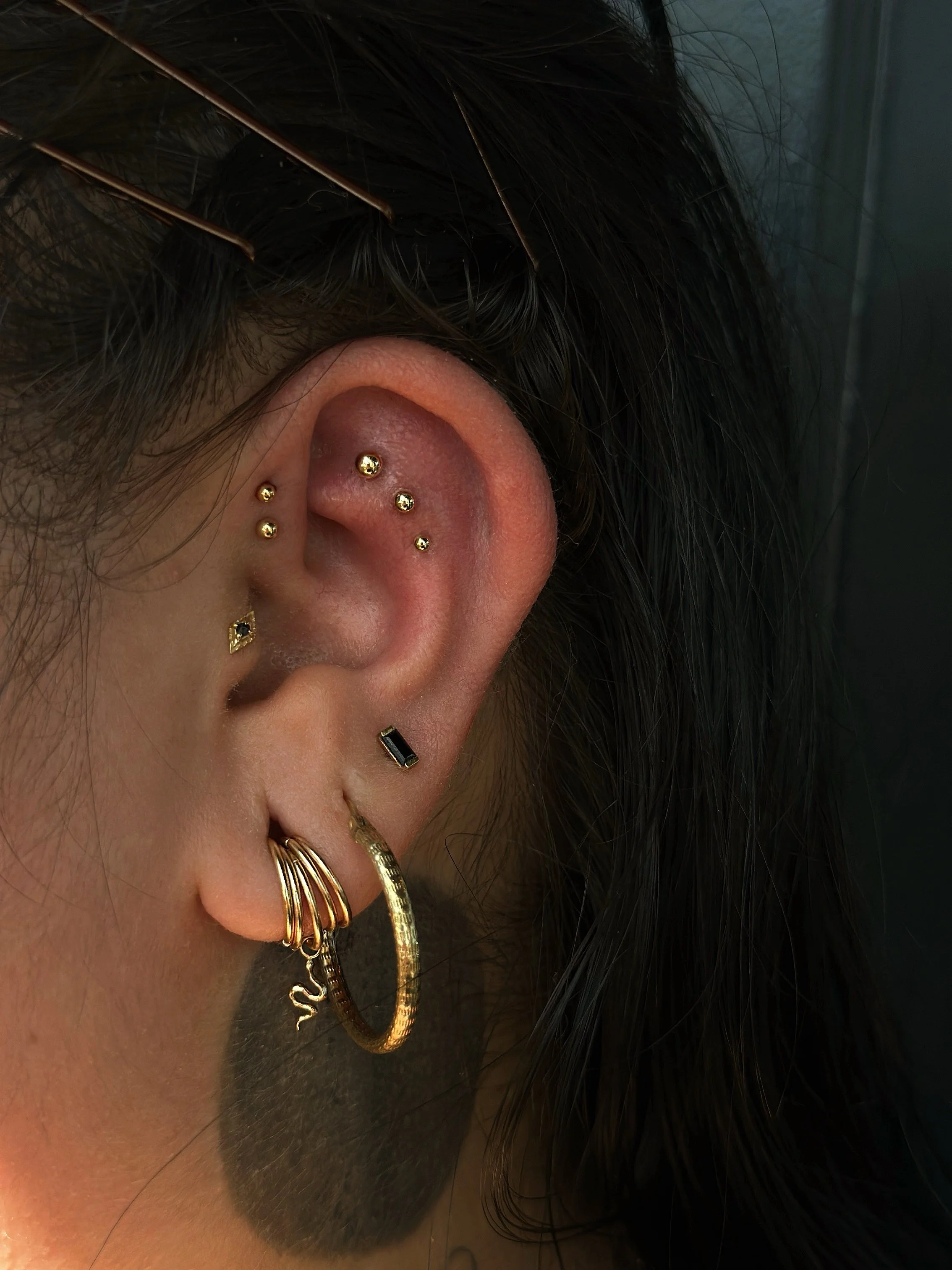 Close-up of a person's ear with multiple gold earrings, including small studs, a black gemstone, a black rectangular earring, and a hoop with a decorative charm, along with dark hair and hairpins.