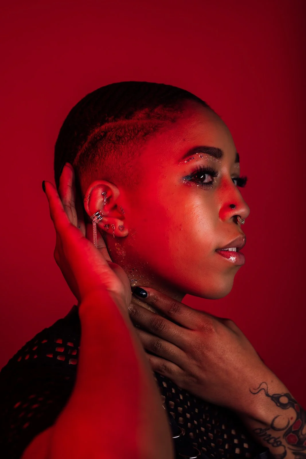 Close-up portrait of a woman with short, shaved hair, multiple earrings, and makeup, against a red background.