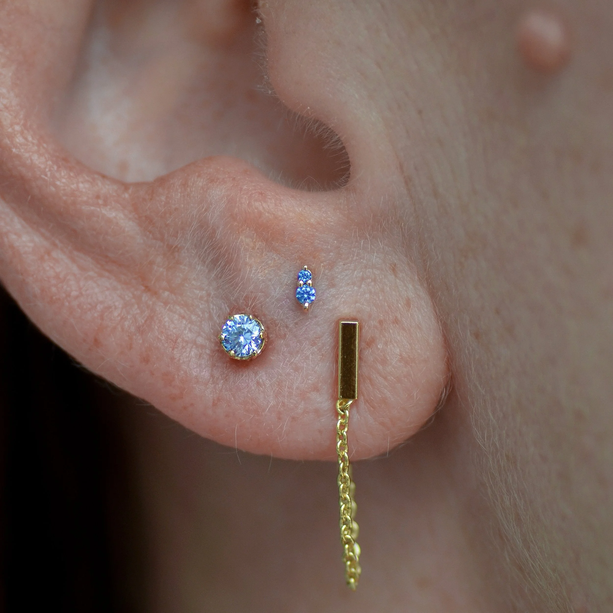 Close-up of a person's ear with three piercings: a large diamond stud, a smaller blue gemstone stud, and a gold dangling chain earring.