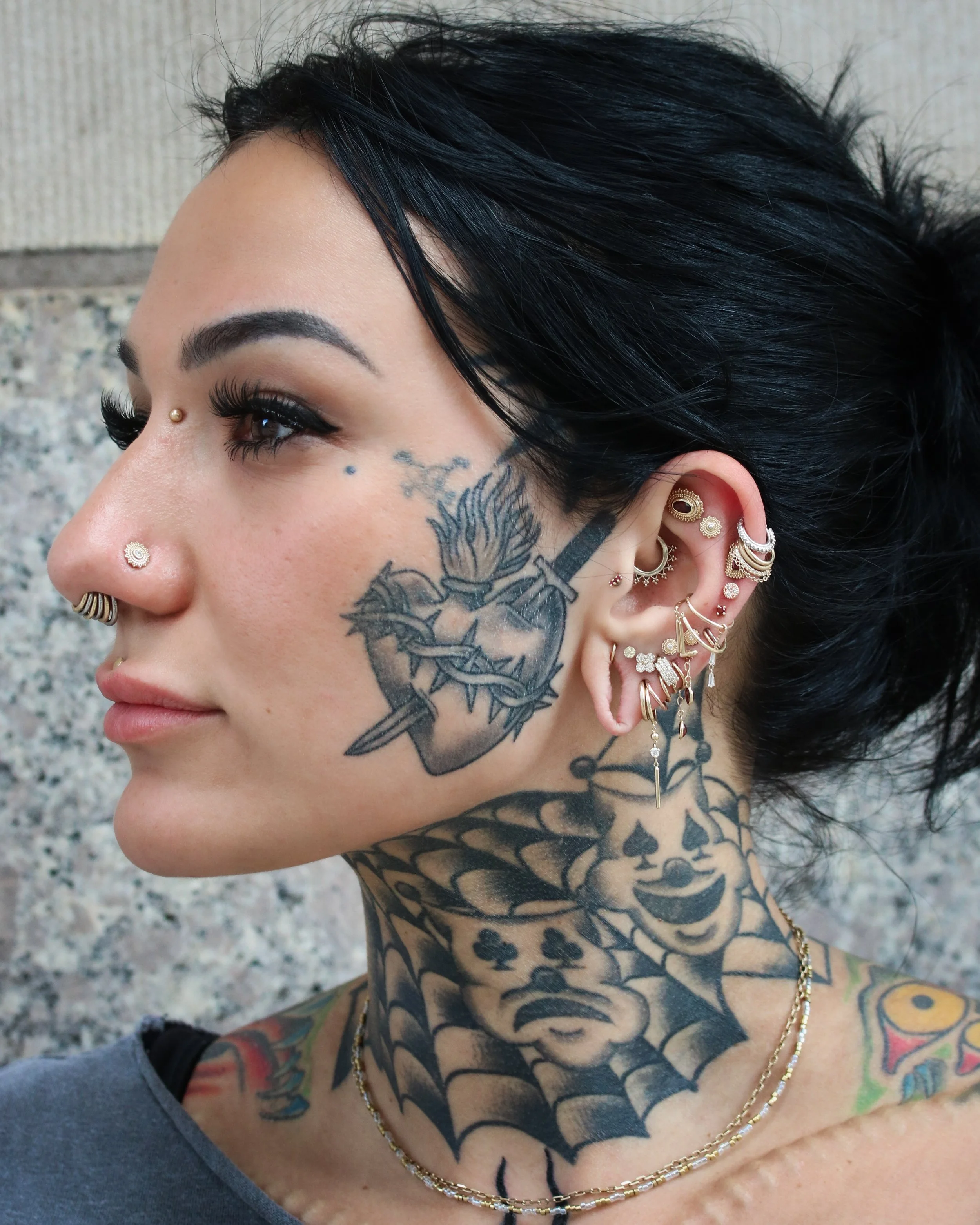 Close-up of a woman with multiple piercings, a tattoo on her face and neck, and tattoos on her chest. She has dark hair and is wearing jewelry.