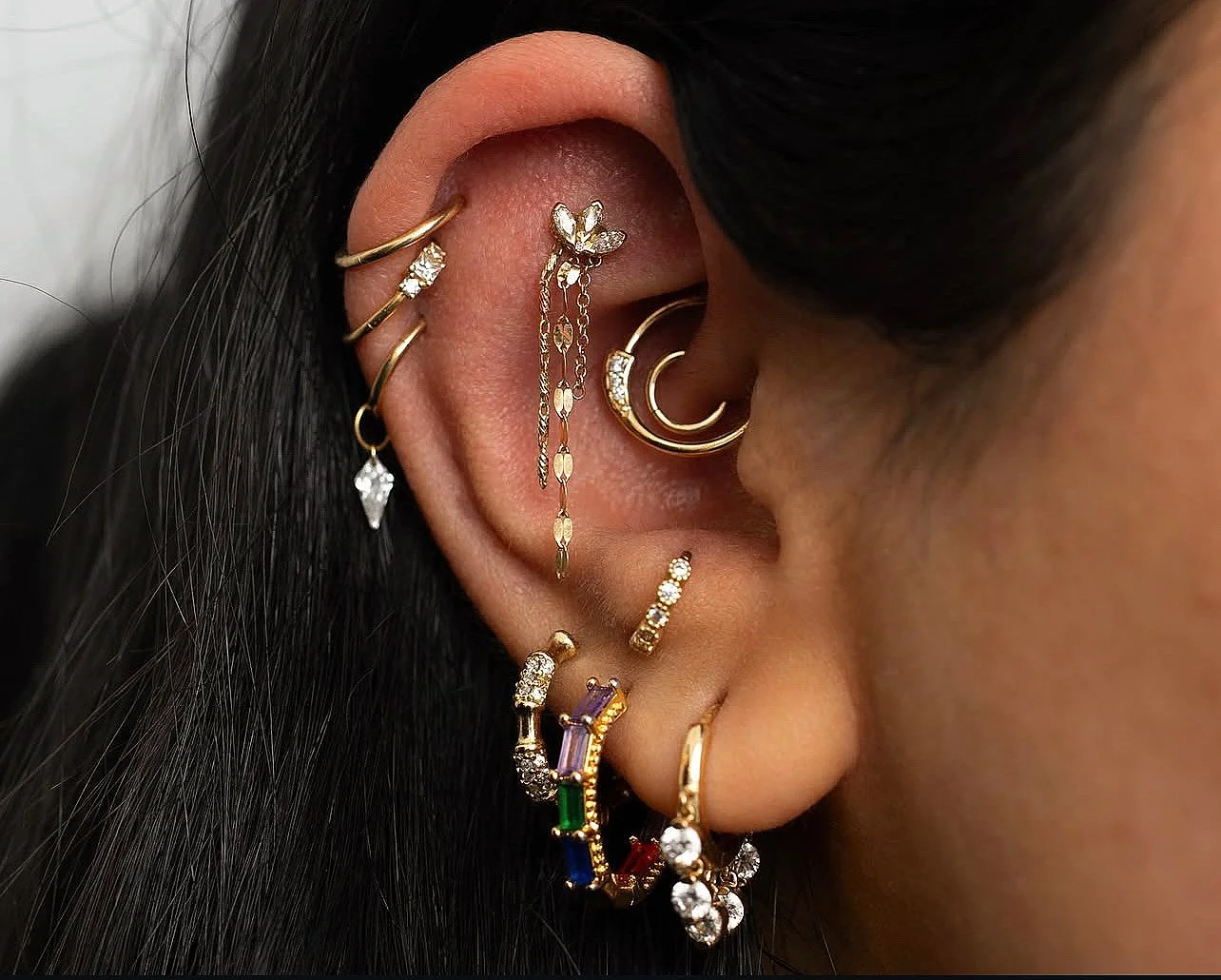 Close-up of a person's ear adorned with multiple gold and diamond earrings, including hoops, chains, and studs, with dark hair partially covering the ear.