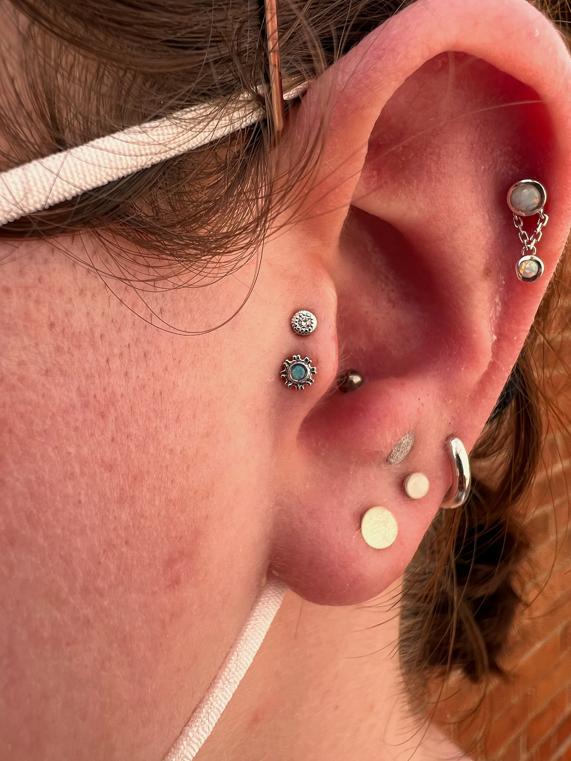 Close-up of a person's ear with multiple piercings, including a helix piercing with a chain-link earring, a rook piercing, a tragus piercing, a lobe piercing, and a second lobe piercing. The person is wearing glasses with a white strap and has brown 